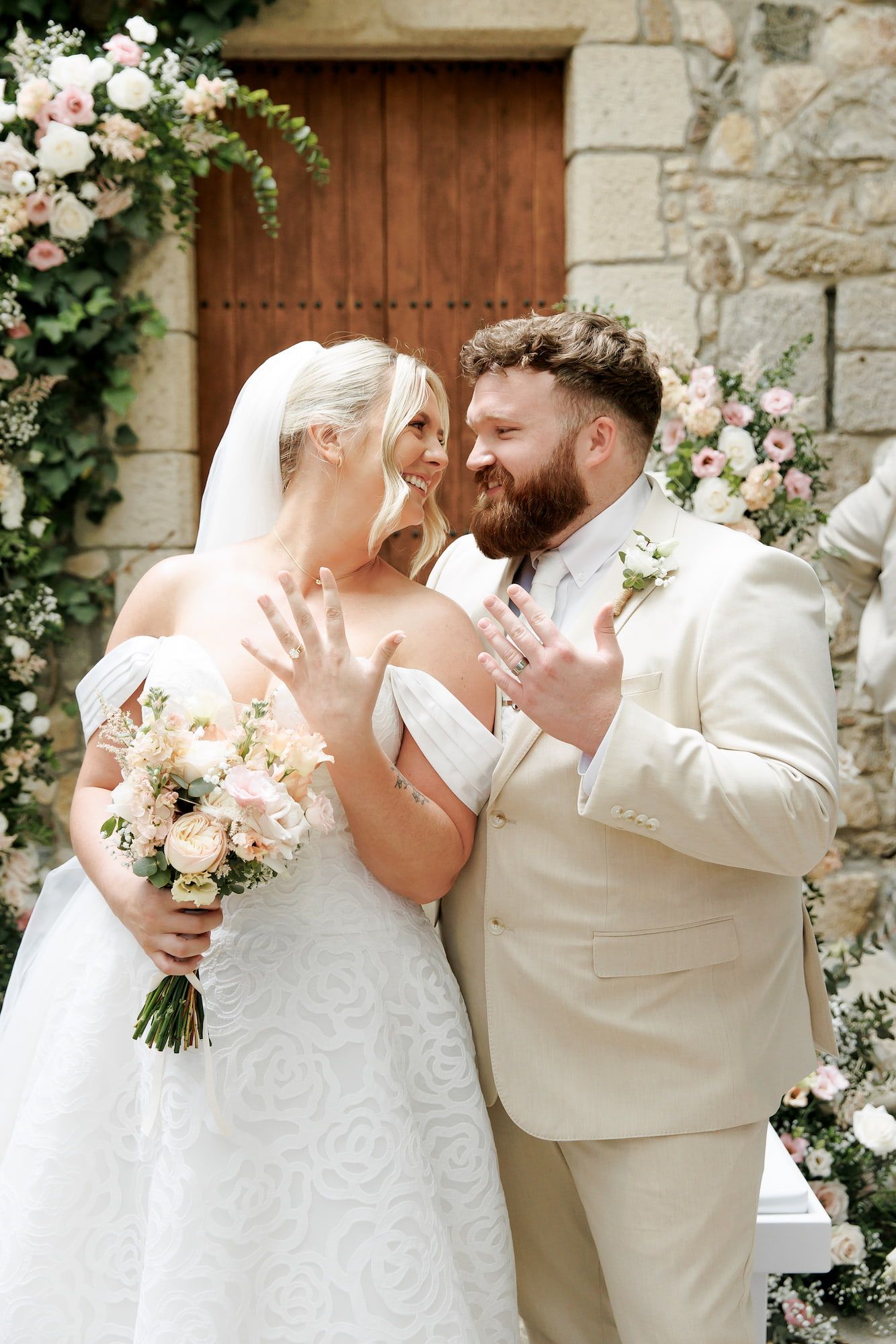 Bride and groom showing wedding rings, smiling at each other, standing before a stone chapel adorned with pink floral arrangements.