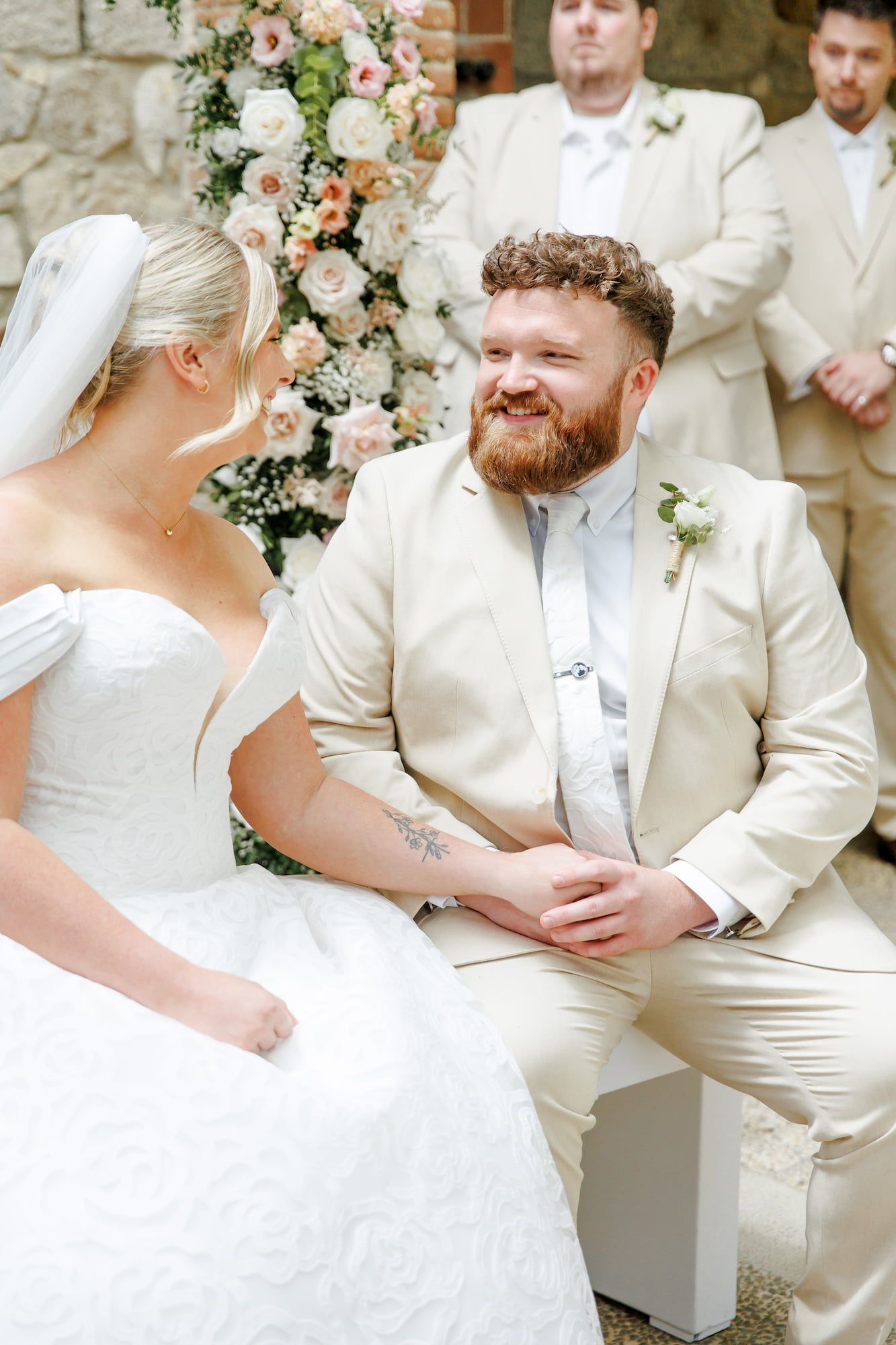 Groom in cream suit smiling at bride in white wedding dress during outdoor ceremony with floral arch.