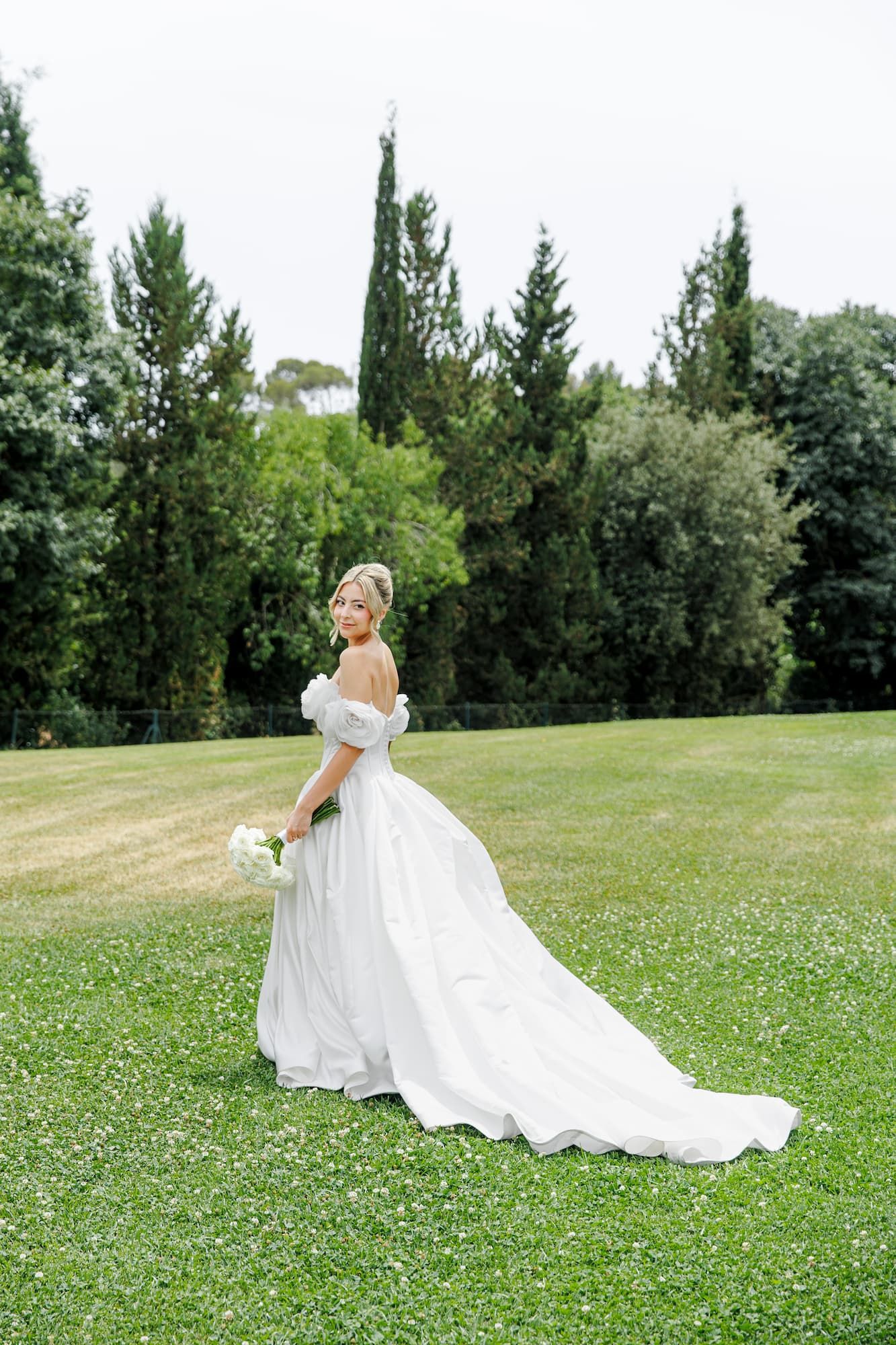 Bride in off-shoulder white ball gown with long train holds bouquet on green lawn with tall trees.