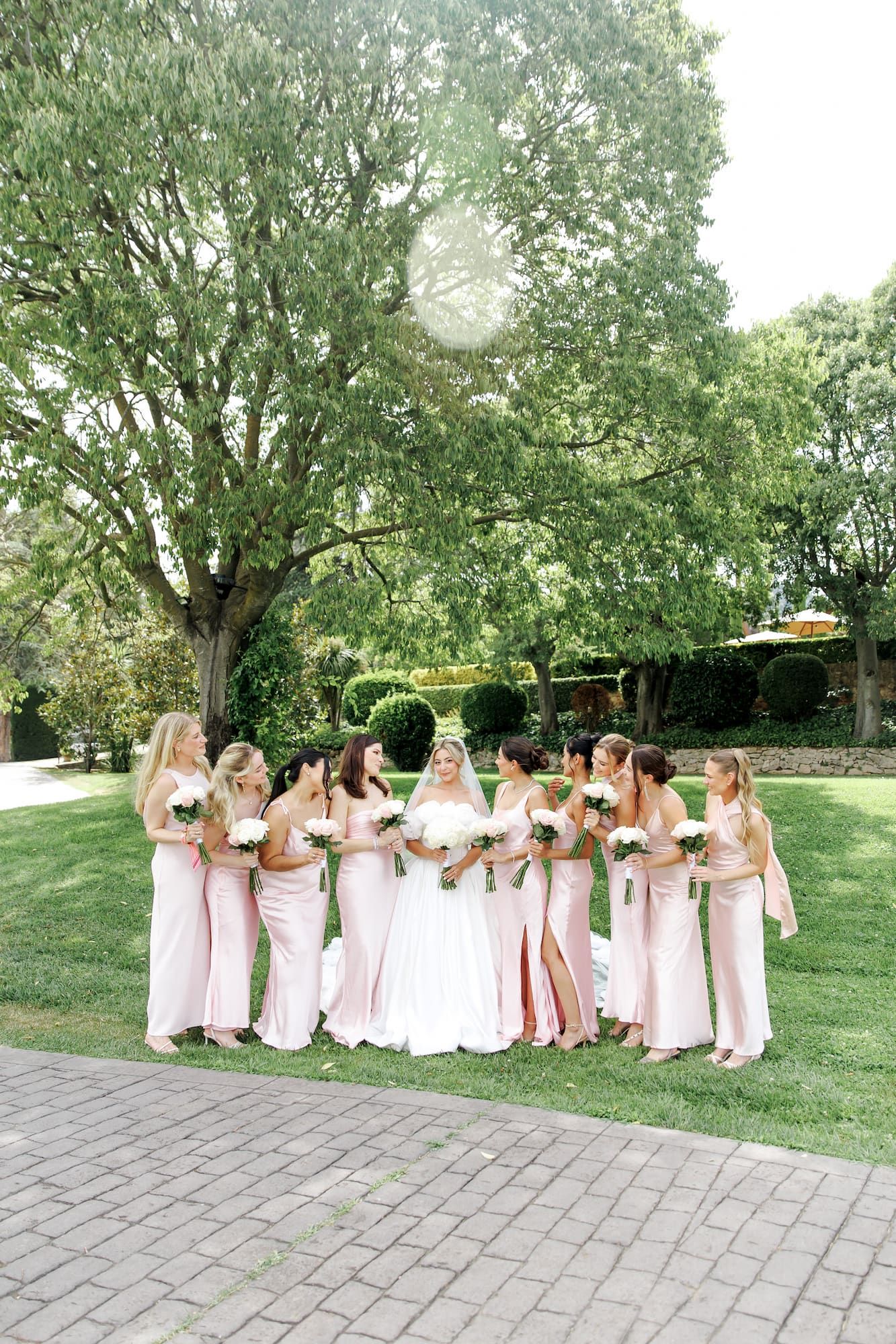 Bride in white gown surrounded by bridesmaids in blush pink dresses holding bouquets on lush green lawn.