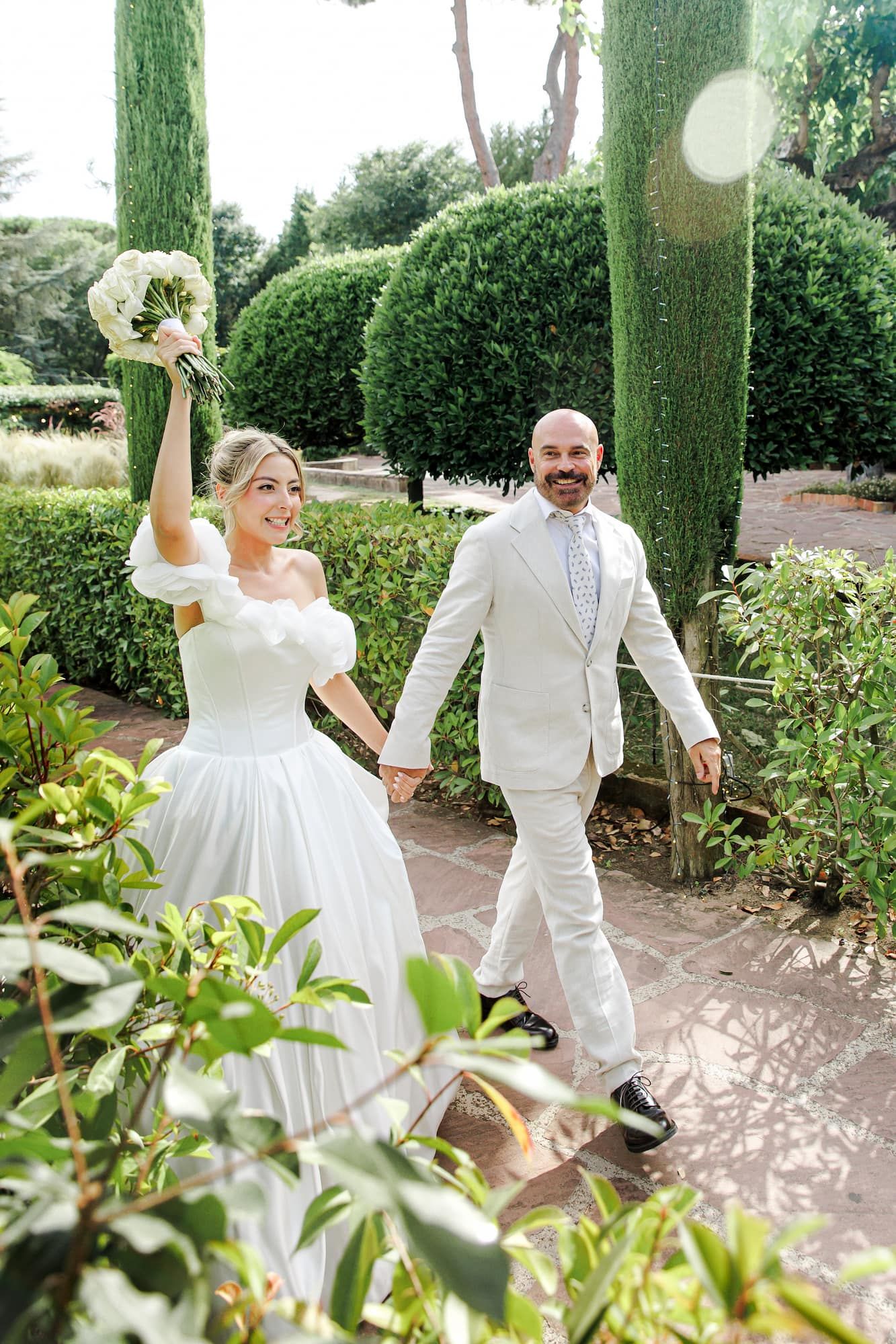 Joyful bride and groom walking hand-in-hand through a lush garden, bride raising bouquet in celebration.