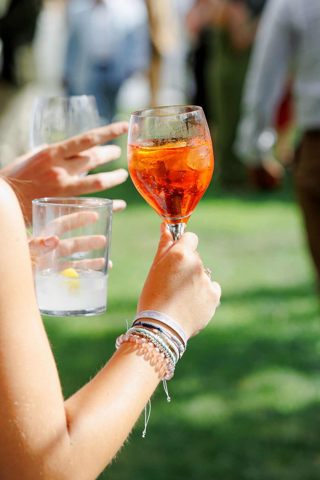 Woman holding an Aperol Spritz cocktail and water glass outdoors at a sunny garden party event.