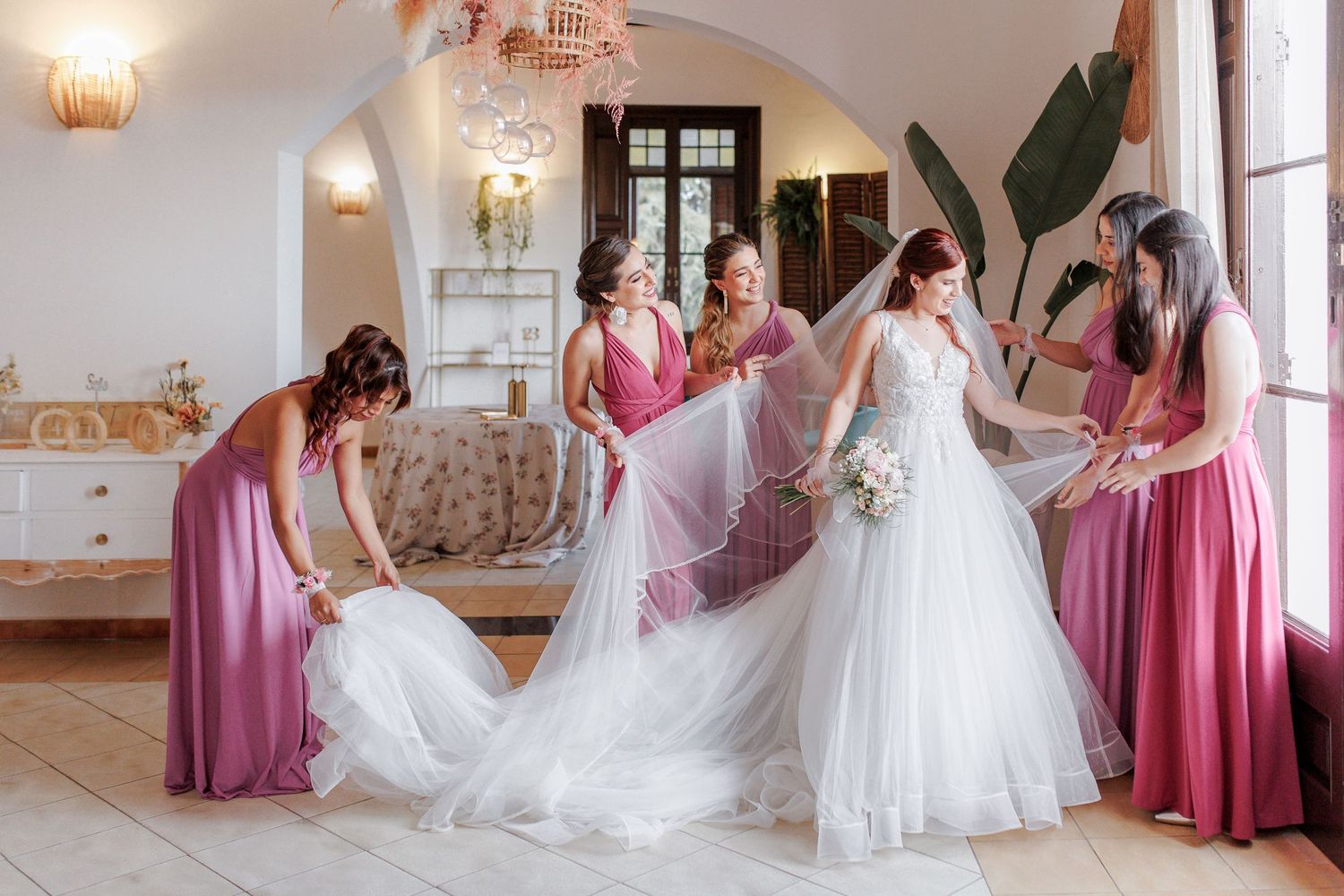 The bride is surrounded by her friends in pink dresses, and they adjust her veil and the designed wedding dress.