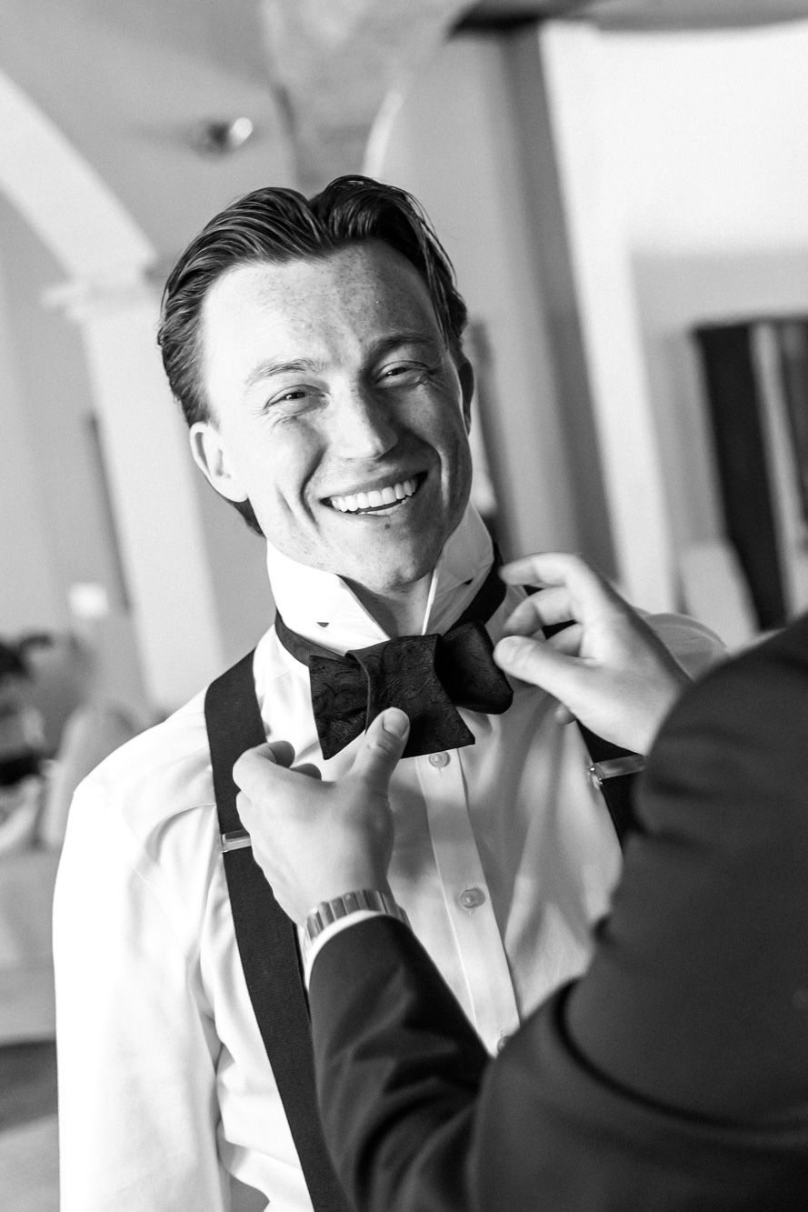 Black and white photo of a smiling groom having his bow tie adjusted by a groomsman before the wedding ceremony.