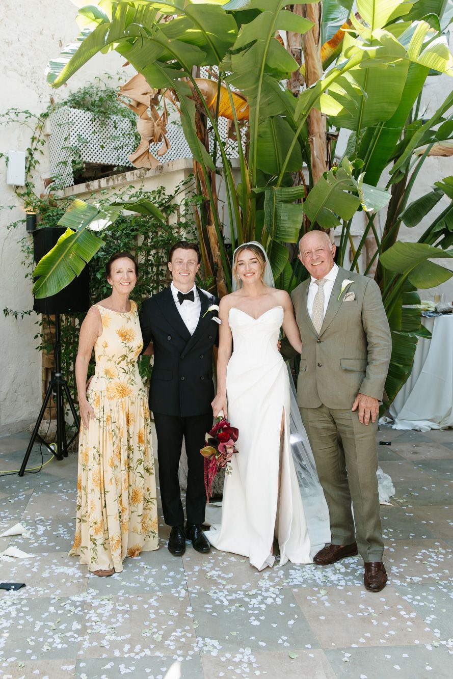 Bride and groom pose with family in wedding attire surrounded by tropical greenery and confetti.