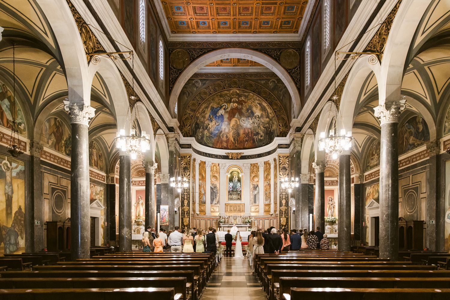 Catholic church interior with ornate frescoes, gilded ceiling, wooden pews, and altar during a wedding ceremony in Barcelona