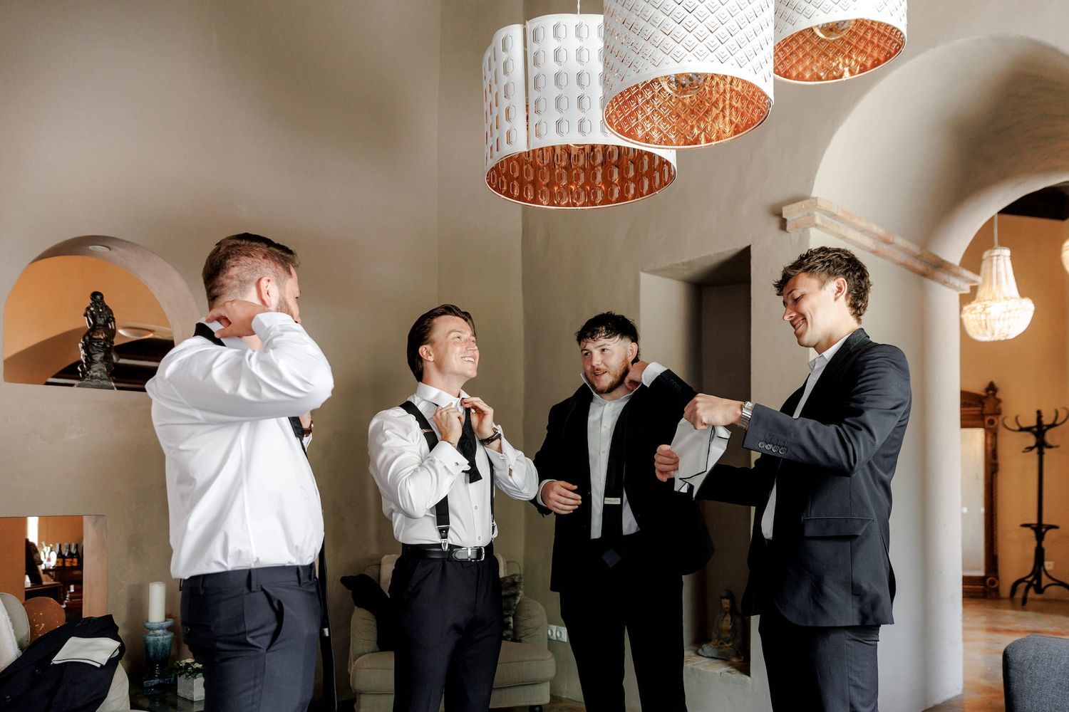 Four groomsmen in black suits and white shirts prepare for a wedding ceremony indoors under decorative pendant lights.