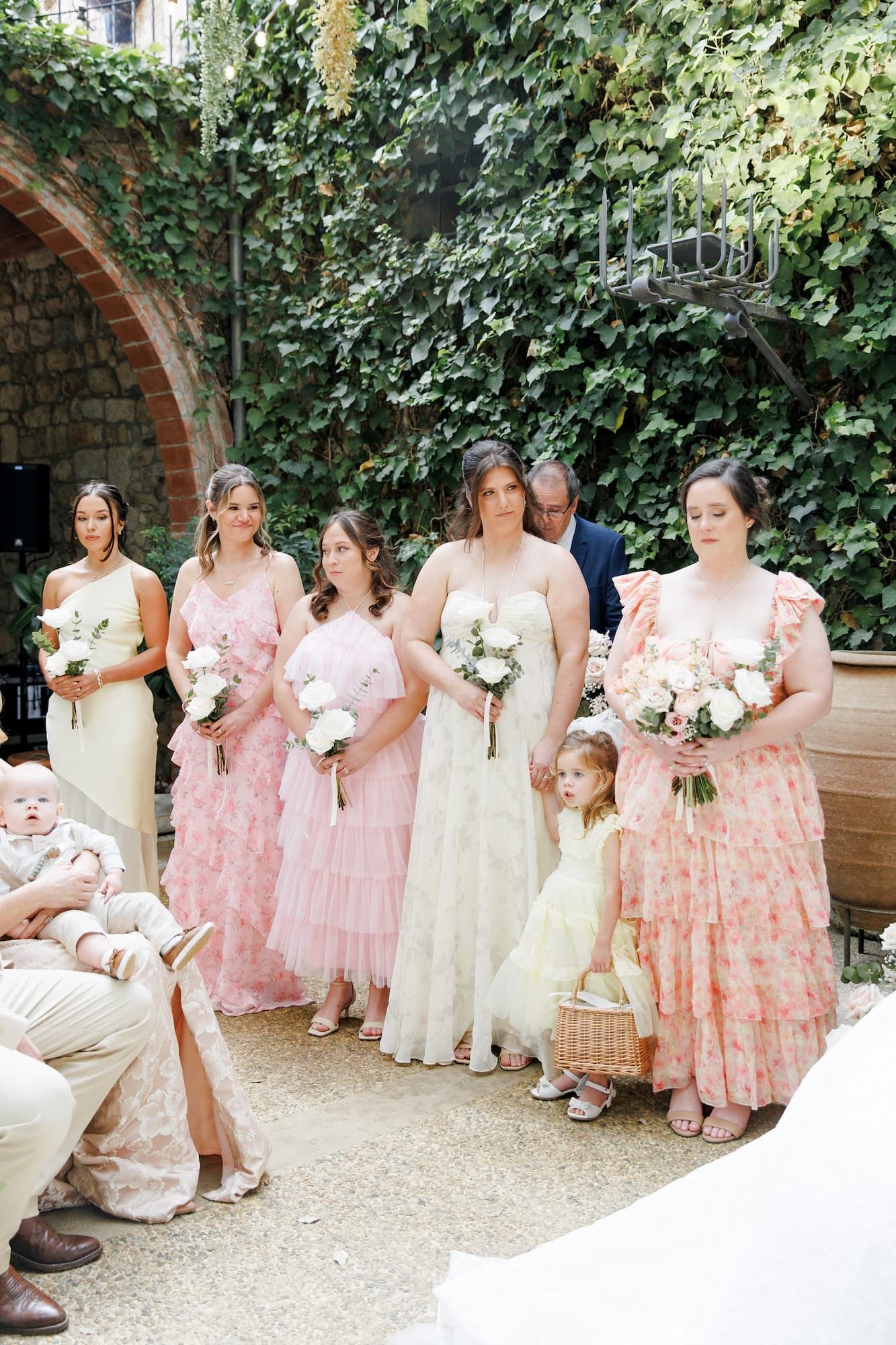 Bride in white gown with bridesmaids in pink dresses and flower girl at outdoor ivy-covered wedding ceremony.