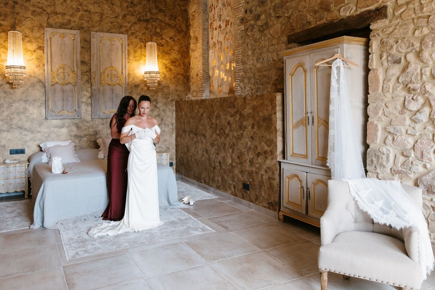 Bride being helped into white wedding gown in rustic stone-walled room with vintage armoire and elegant decor.