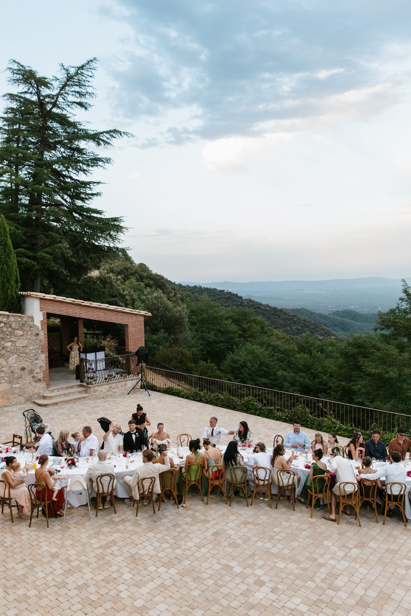 Outdoor wedding reception on a terrace with mountain views, guests seated at white-clothed tables surrounded by lush greenery.