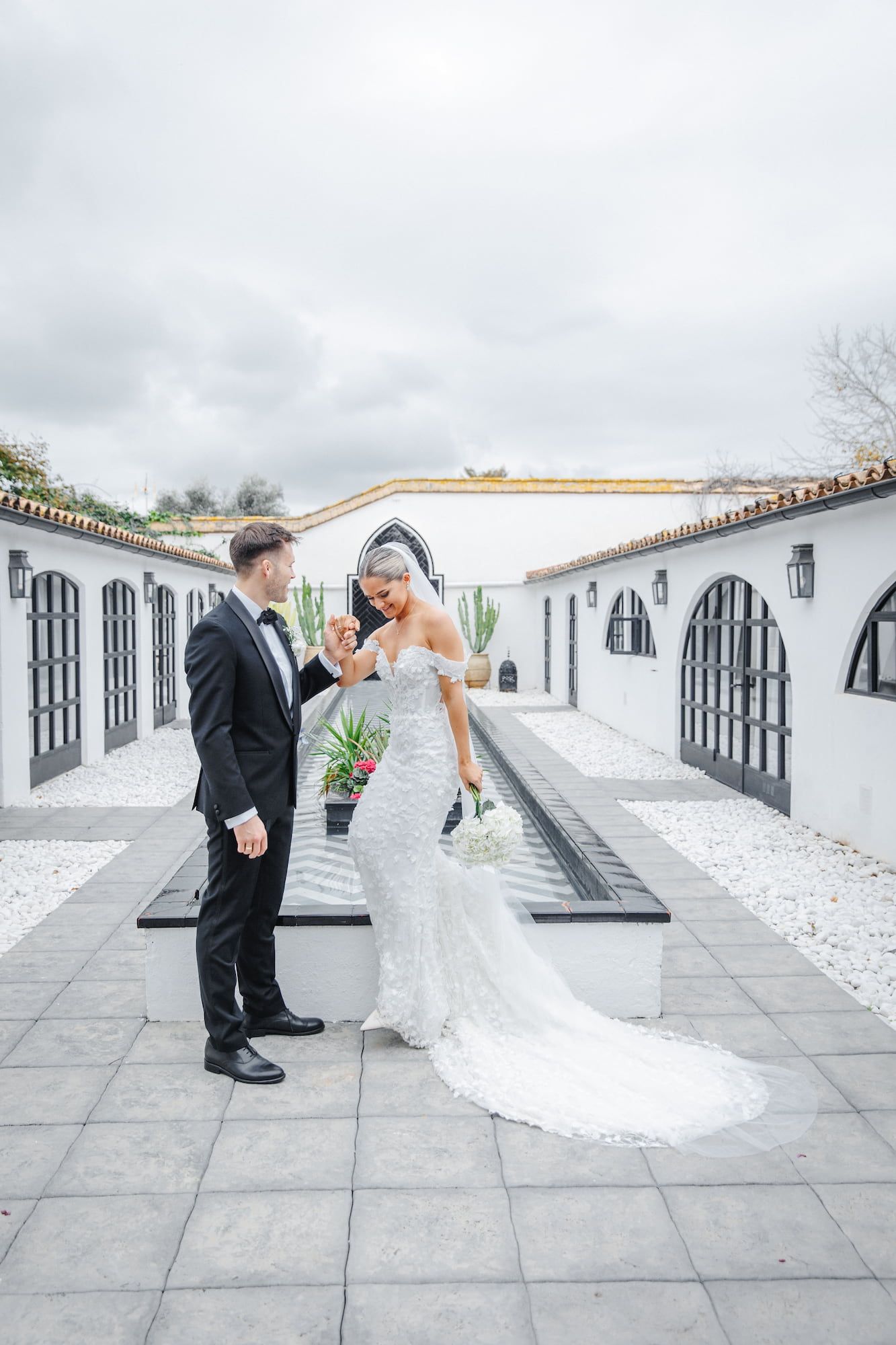 Bride and groom share a romantic moment in a white Spanish-style courtyard, she in a lace gown, he in a black tuxedo.