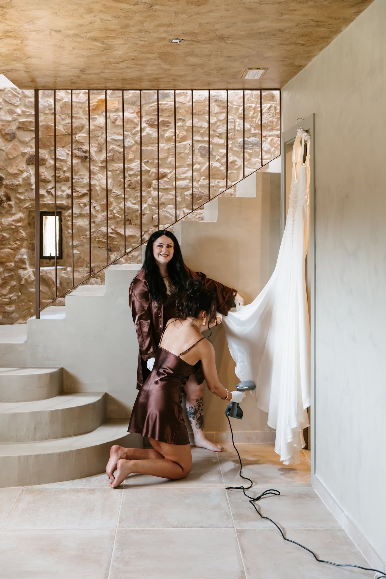 Bride in brown silk robe getting dress steamed by bridesmaid in rustic stone staircase setting.