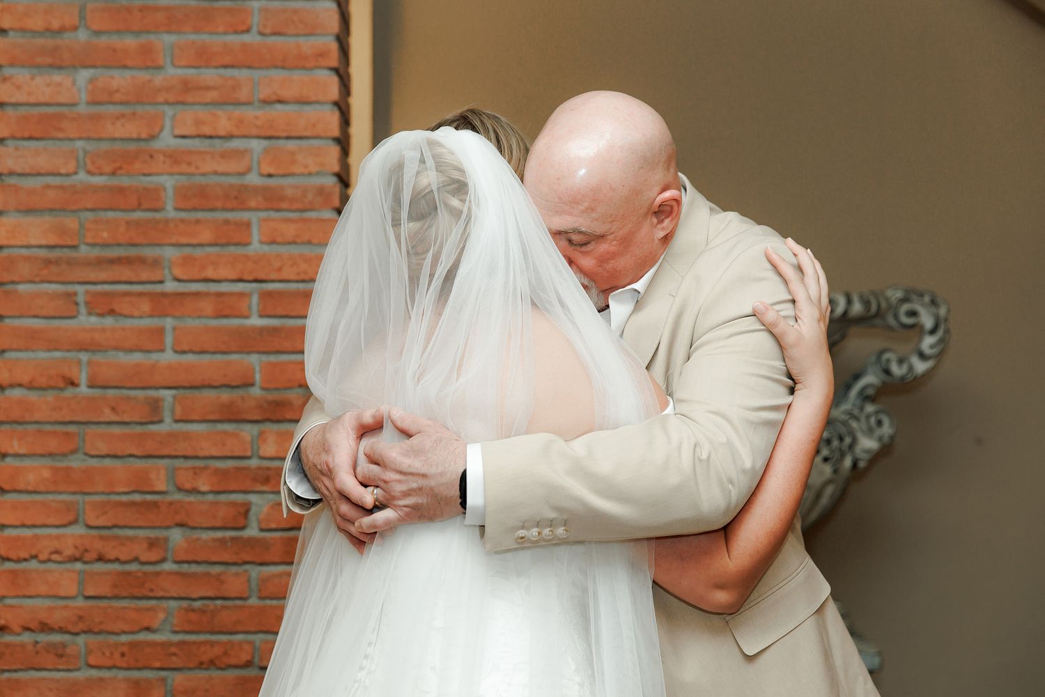 Bald groom in beige suit embracing bride in white wedding dress and veil near brick wall.
