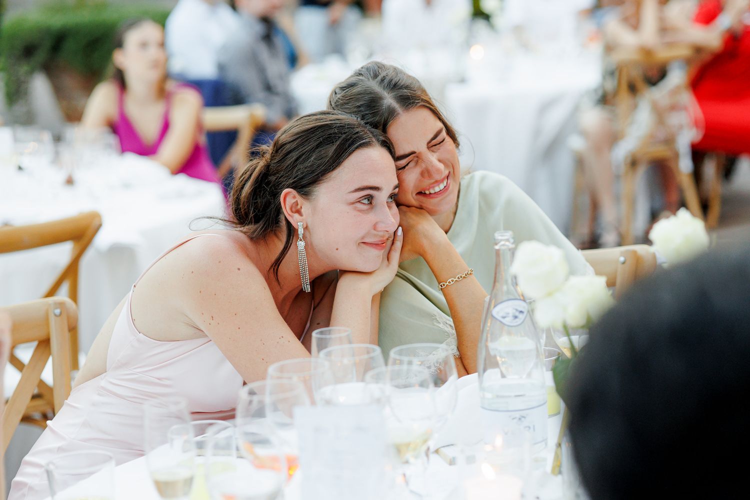 Two women lean together smiling at an outdoor wedding reception table with white flowers and glassware.