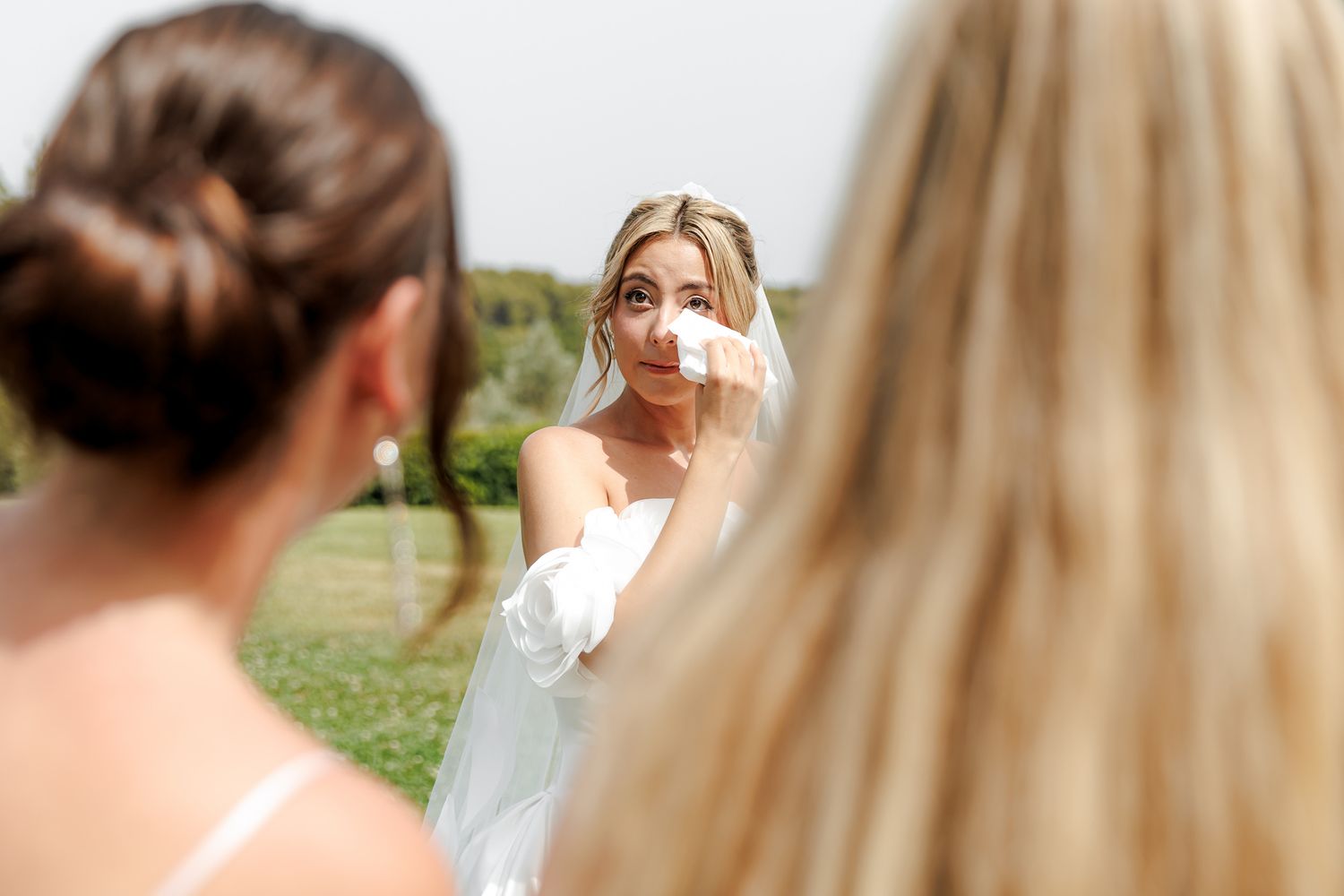 Emotional bride in white gown and veil wiping tears during wedding day first look with bridesmaids.
