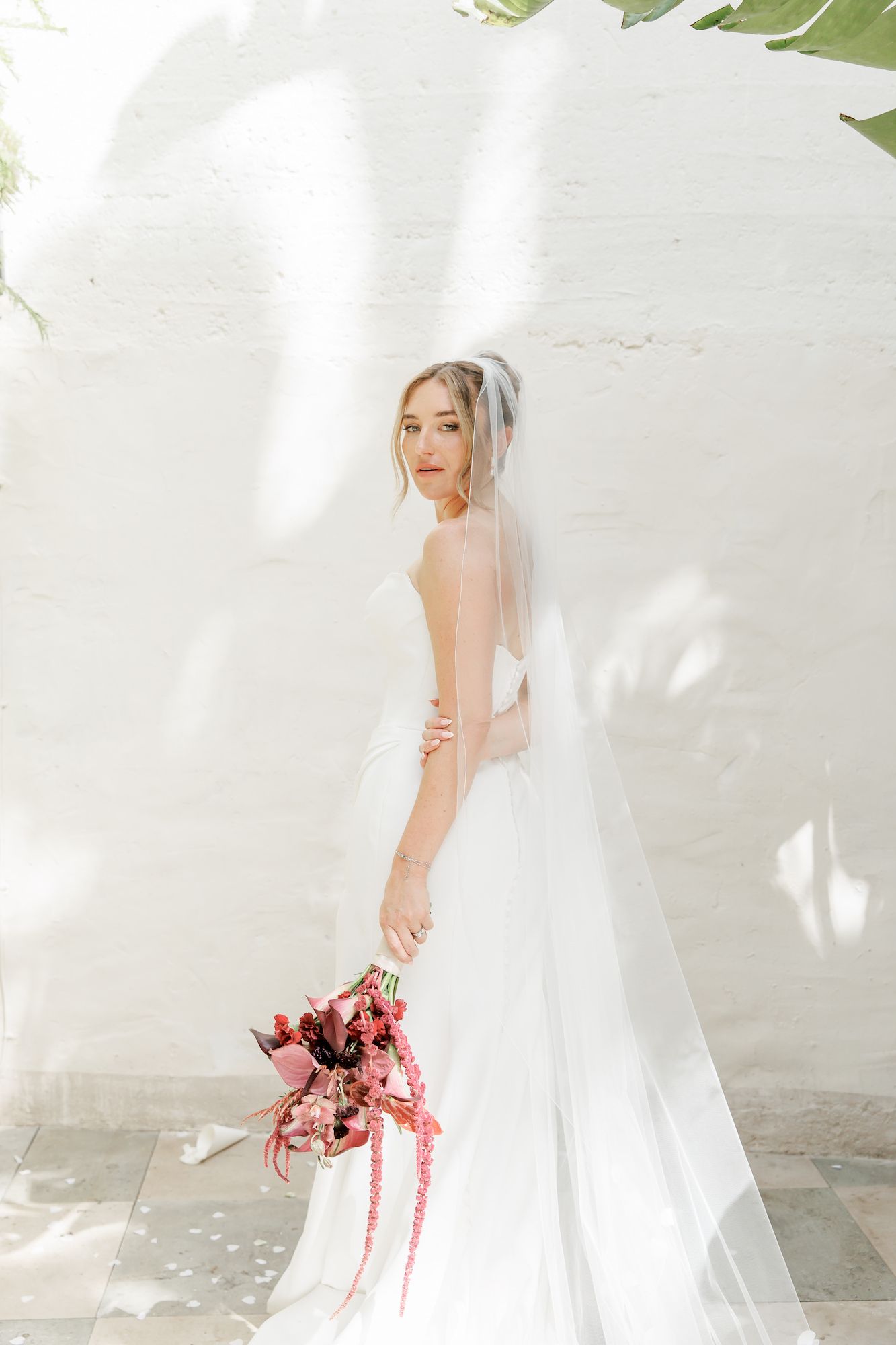 Bride in white gown and veil holds pink bouquet against sunlit white brick wall.