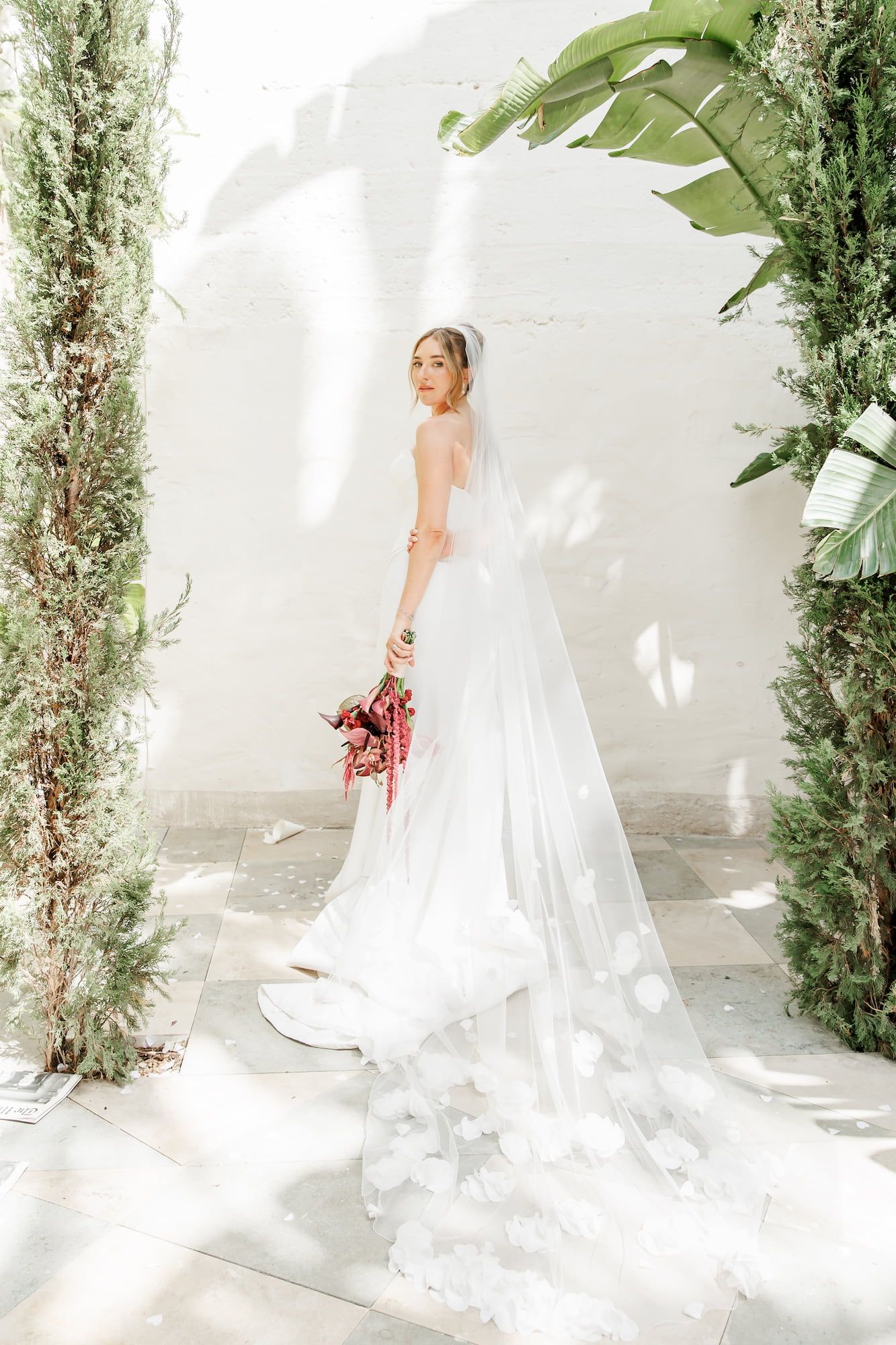 Bride in a white gown and a floral veil holds a red bouquet, standing between lush greenery in sunlit garden at a wedding venue in Barcelona