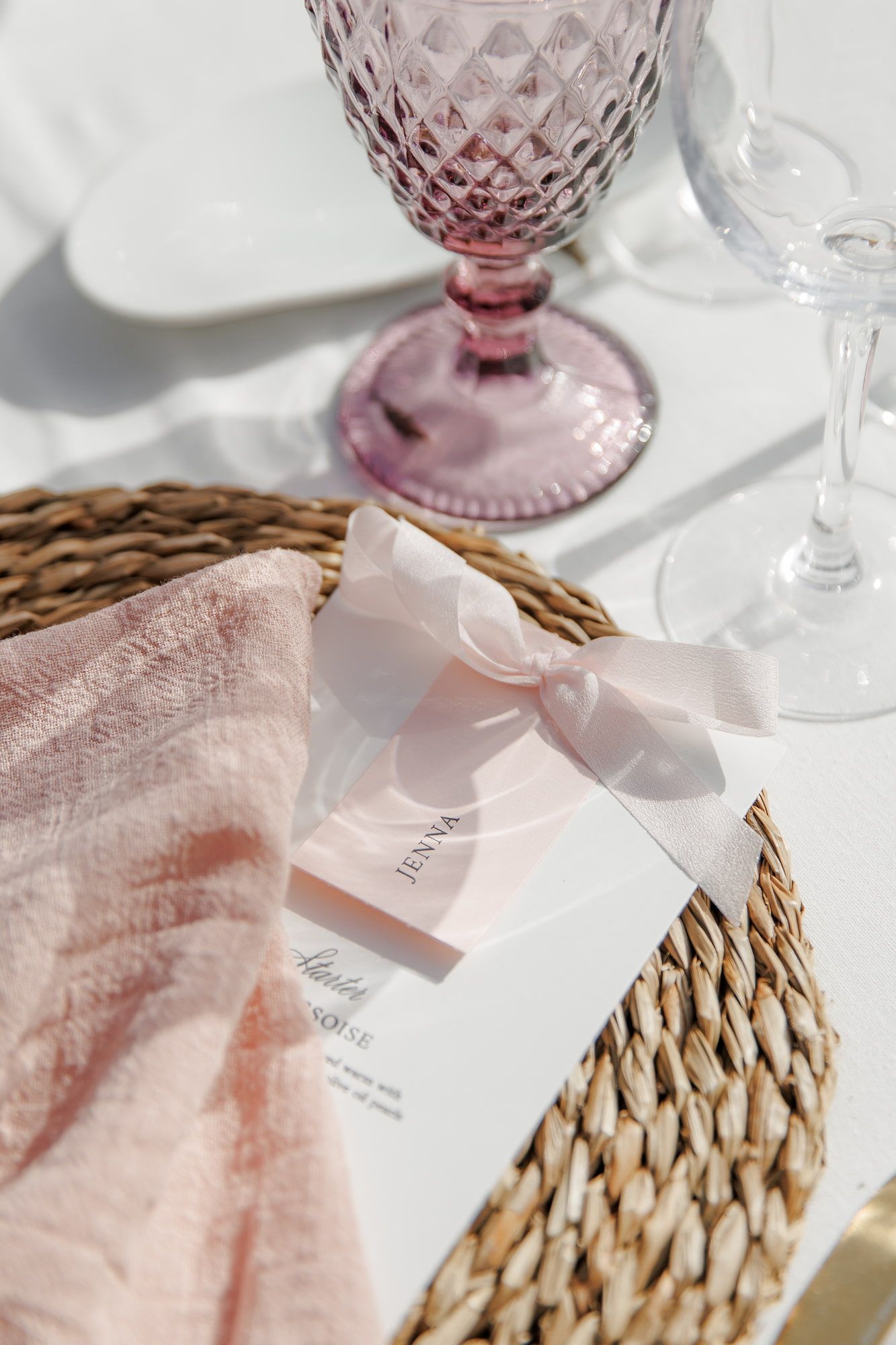Elegant wedding place setting with pink napkin, ribbon-tied menu card, and vintage pink goblet on wicker charger.