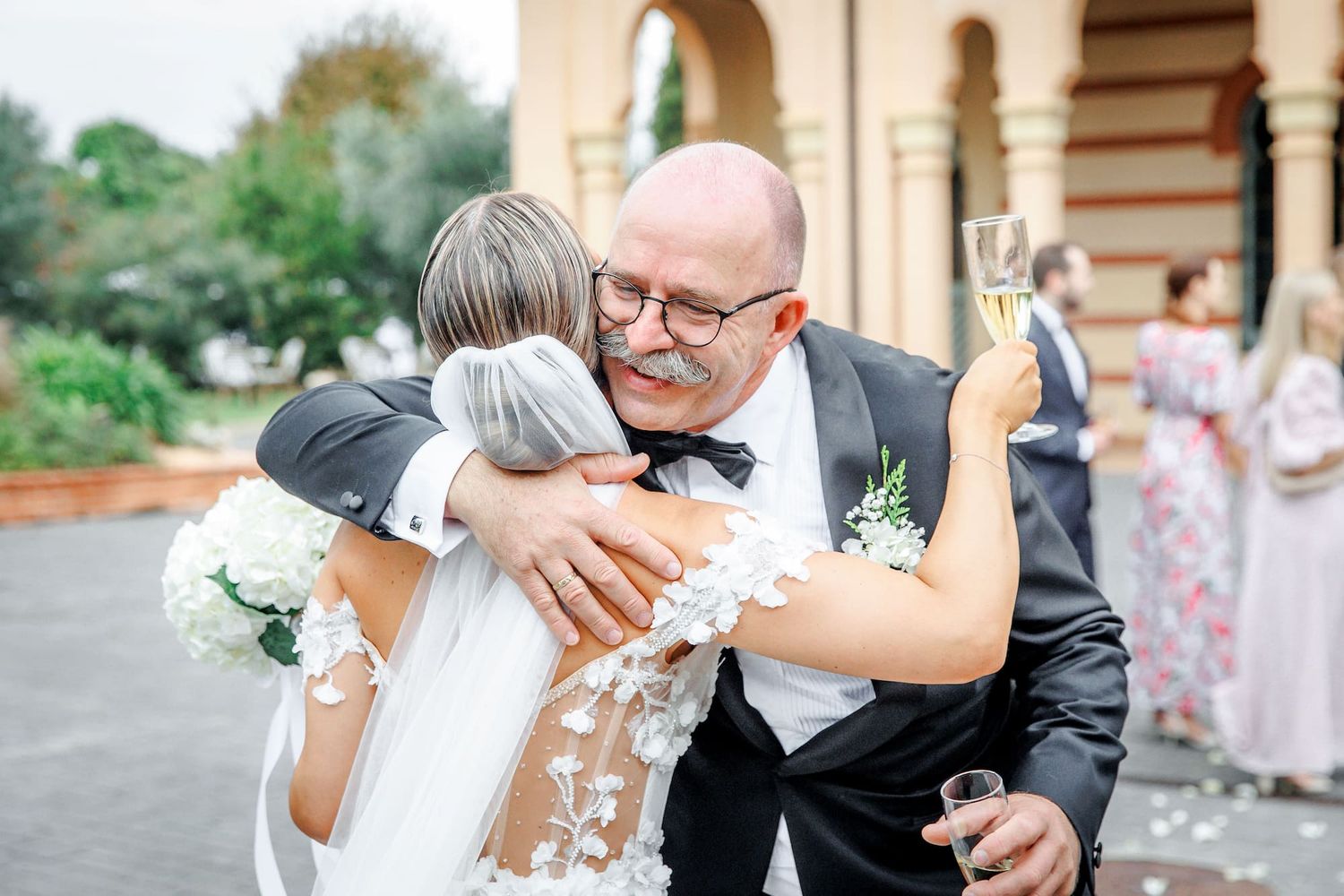 Bald man in tuxedo warmly embracing bride in lace gown at elegant outdoor wedding celebration in Barcelona