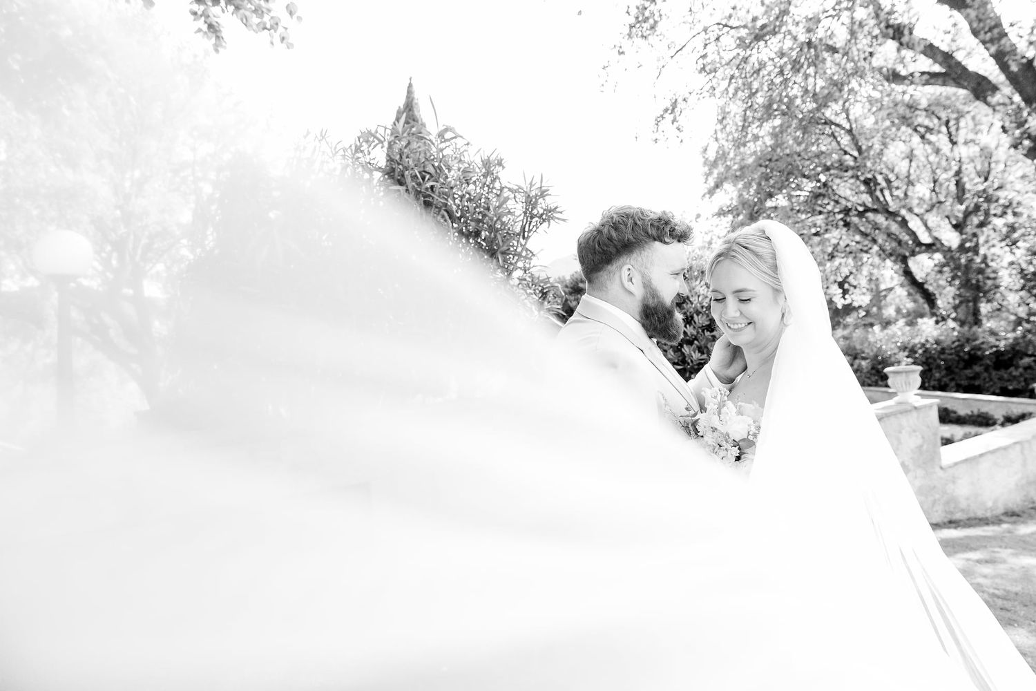Black and white wedding photo of bride and groom embracing, with flowing veil billowing around them outdoors.