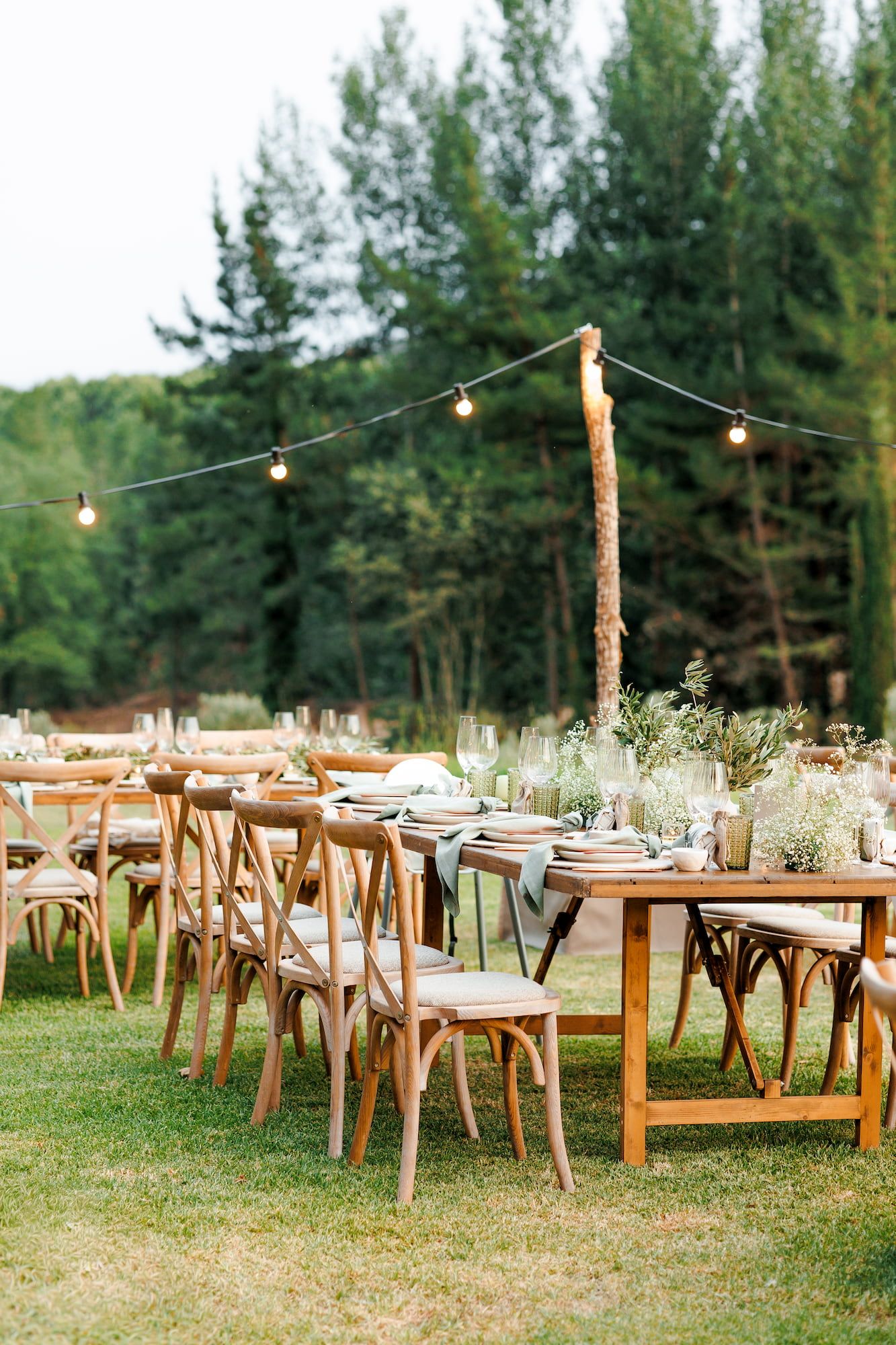 Outdoor wedding reception with rustic wooden farm tables, cross-back chairs, floral centerpieces, and string lights among tall trees.
