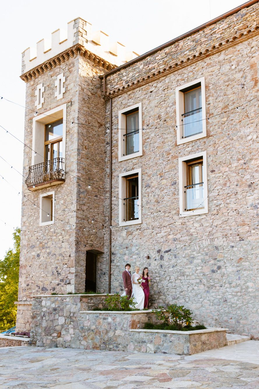 Bride in a white gown and groom in a burgundy suit pose on stone steps beside a castle-style stone building with a crenellated tower.