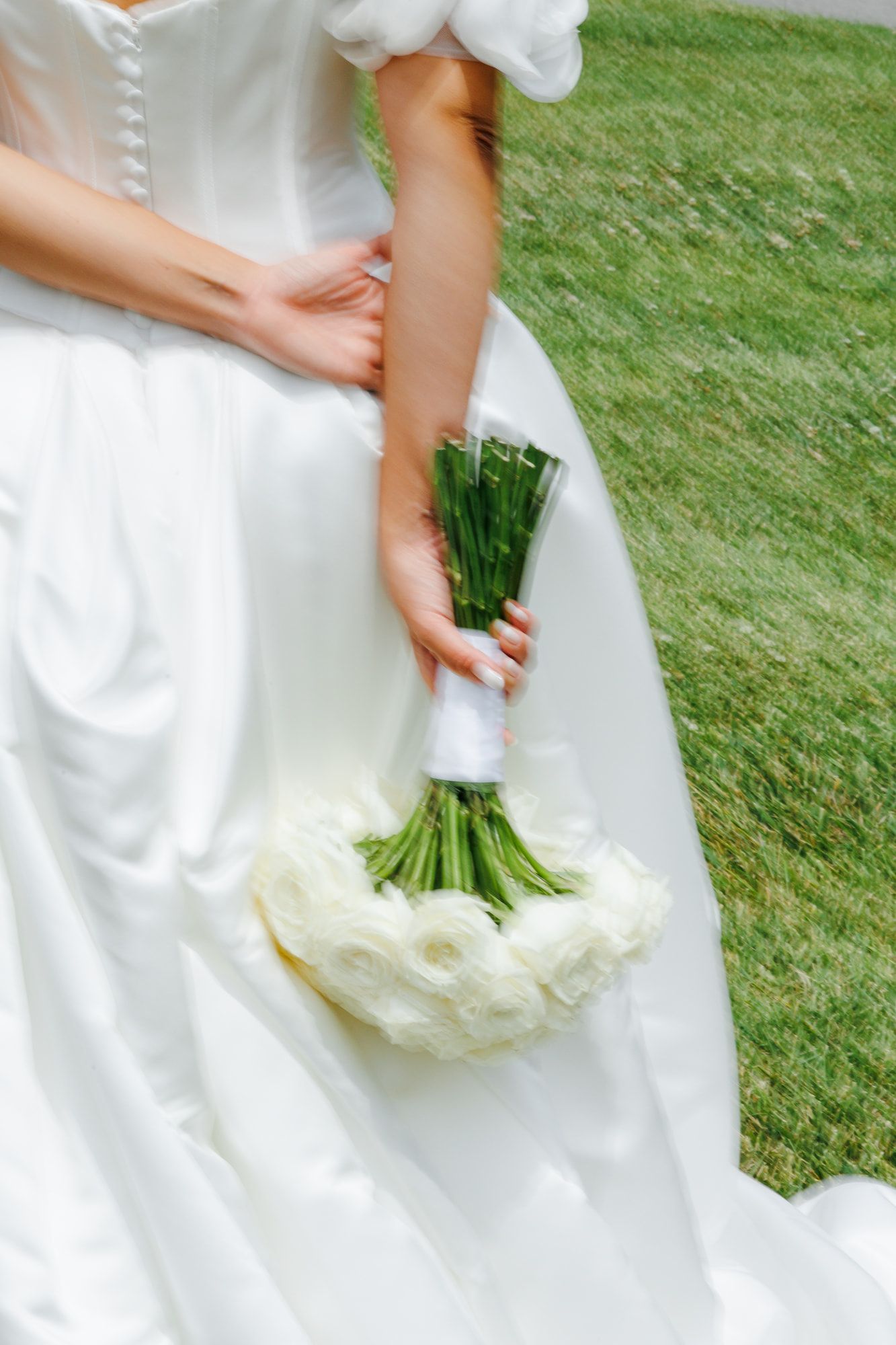 Bride in white wedding gown holding elegant white rose bouquet with green stems wrapped in ribbon on lush grass.