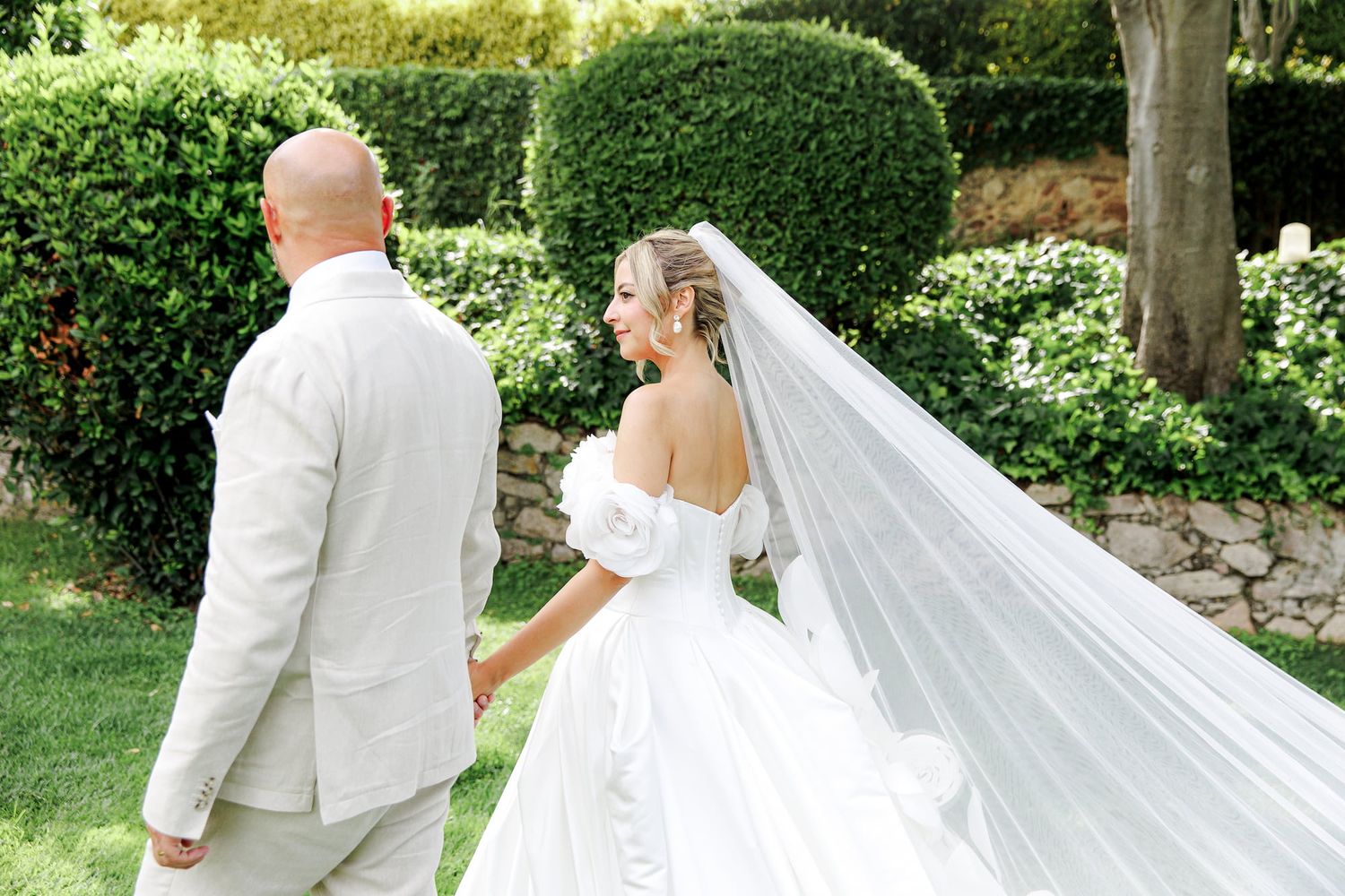 Bride in off-shoulder gown with flowing veil holds hands with bald groom in tan suit walking through garden.