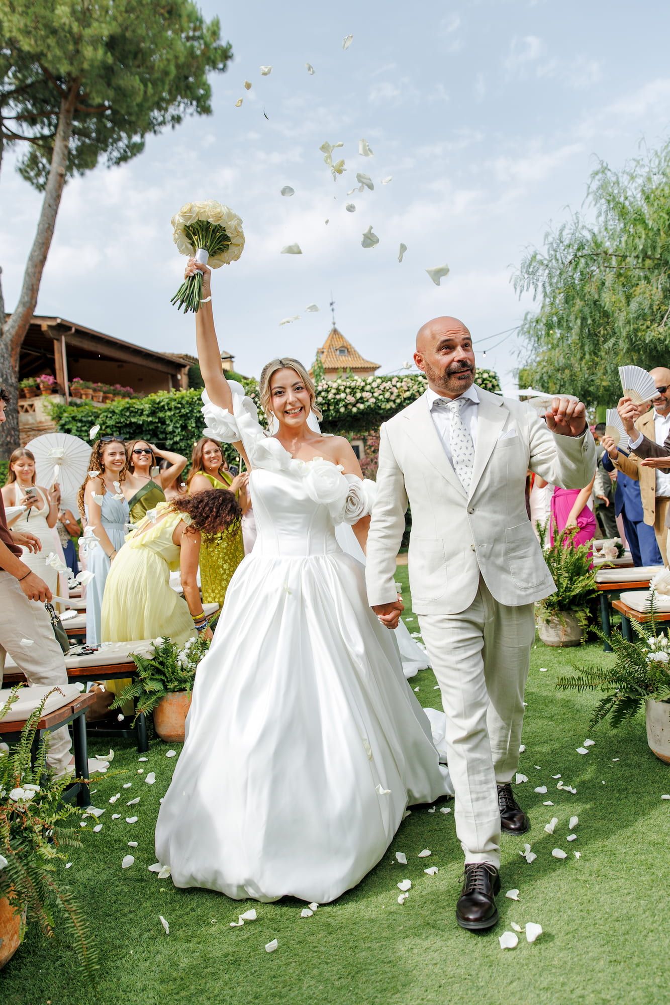 Bride and groom walk down the aisle as guests toss confetti at an outdoor wedding ceremony.
