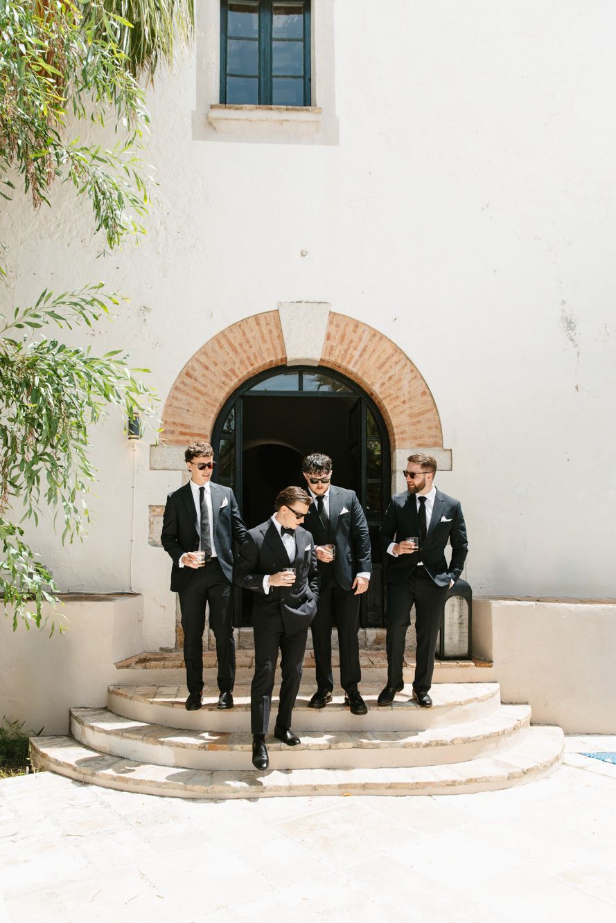 Groomsmen in black suits with sunglasses descend stone steps outside a white building with arched doorway.