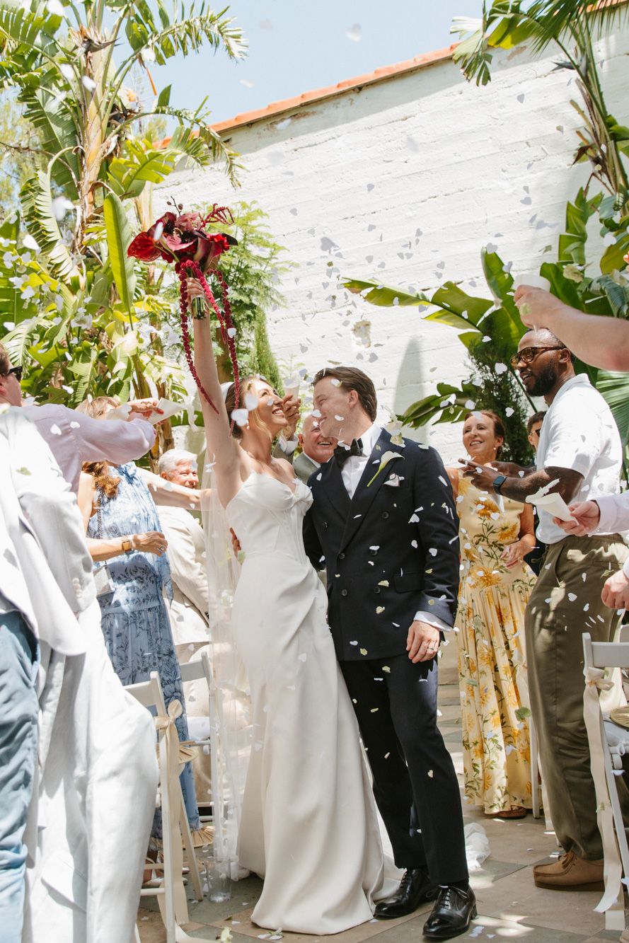 Bride and groom kiss during confetti exit at outdoor wedding ceremony surrounded by cheering guests at Masia Victoria, Barcelona