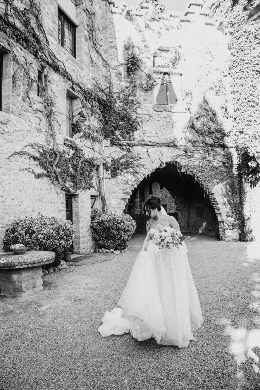 Black and white photo of a bride in a flowing gown holding a bouquet outside a stone castle venue.
