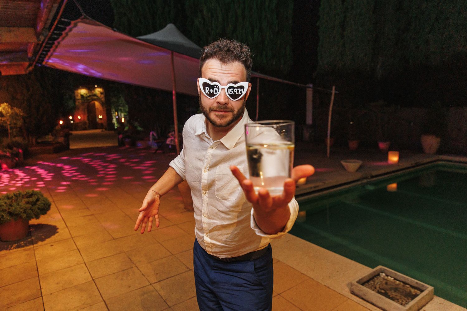 Man in heart-shaped sunglasses holding a drink at an outdoor pool party at La Baronia