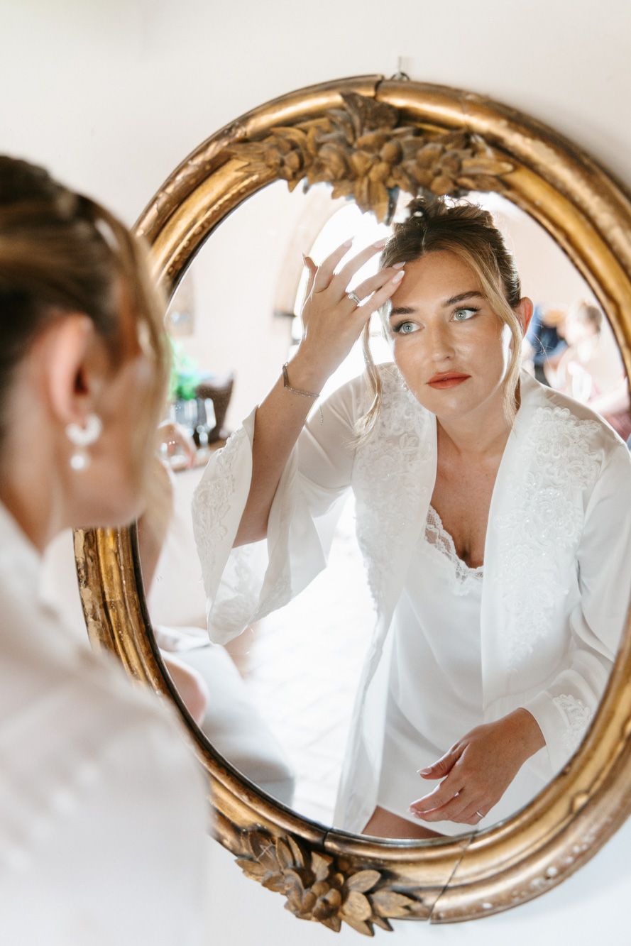 Bride in white robe checks hair in a gold oval mirror, wearing pearl drop earrings and floral hair accessory.