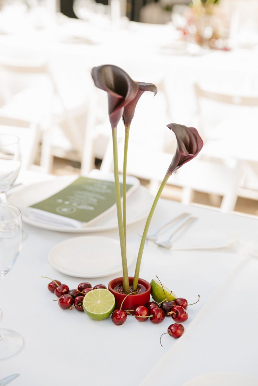 Elegant wedding table centerpiece with dark calla lilies in red pot, surrounded by fresh cherries and lime halves.