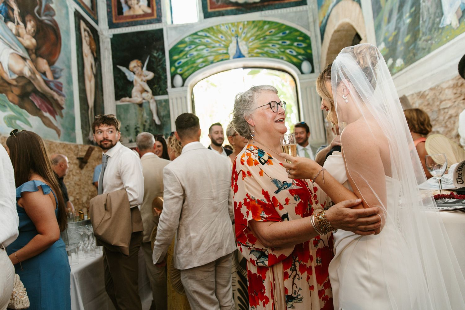 Elderly woman in floral dress smiling at bride in white veil during wedding reception in ornate chapel.