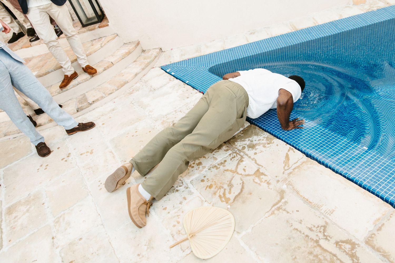 Man lying face down on tiled pool deck beside a swimming pool while others stand nearby.