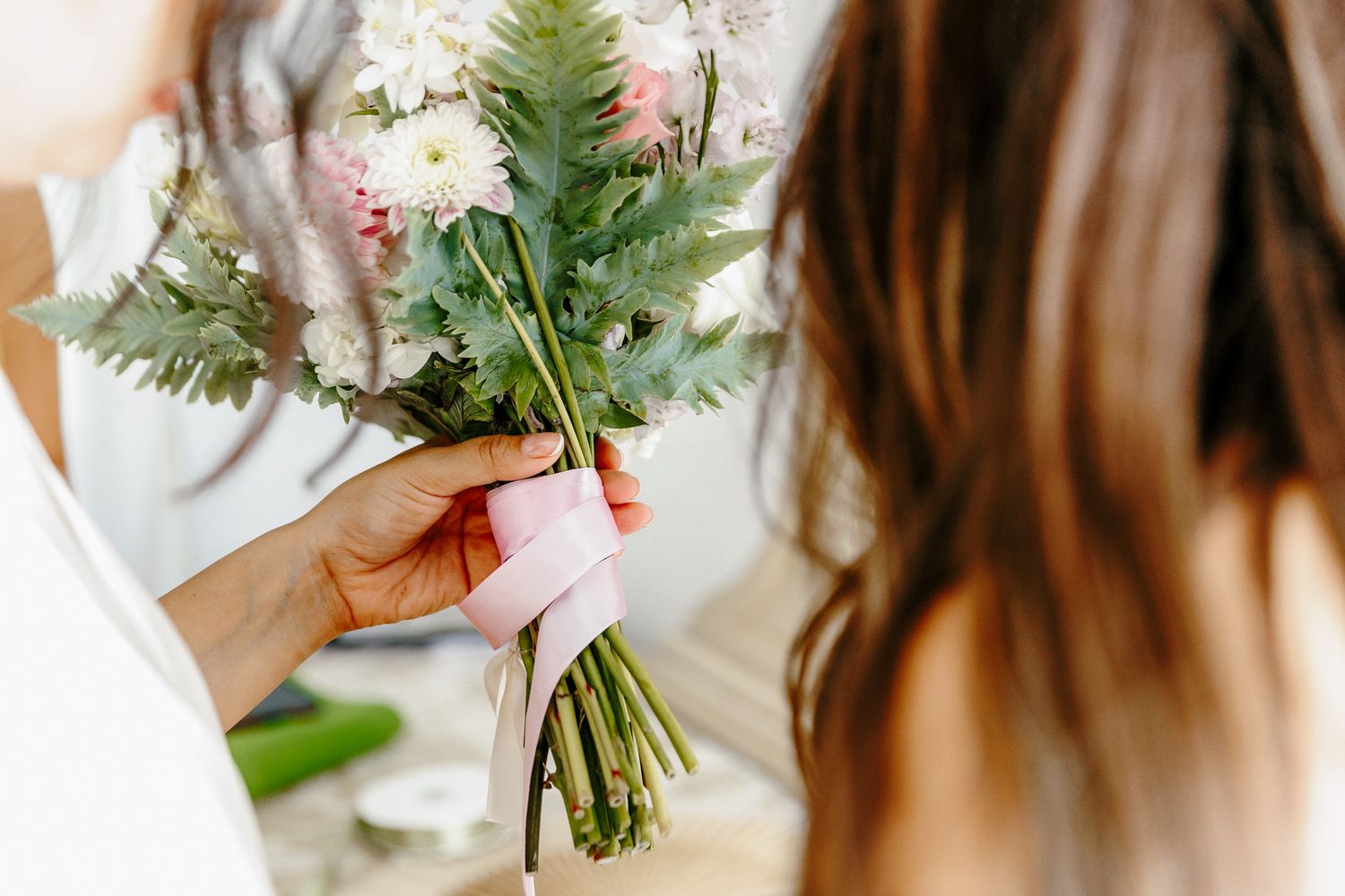 A hand holds a delicate bridal bouquet of white flowers and ferns tied with pink ribbon near a bride.