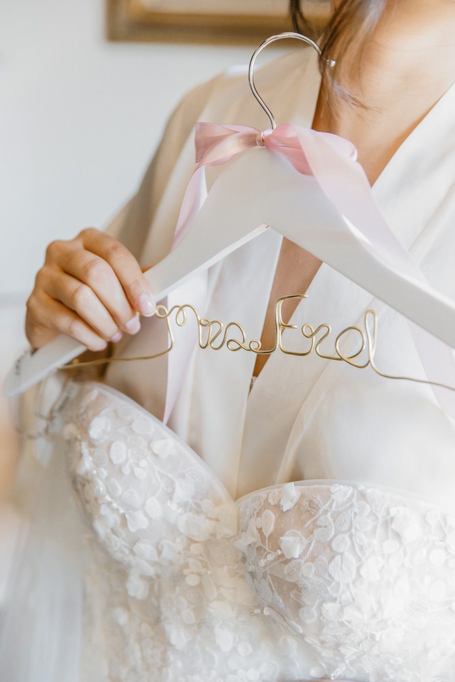 Bride holding a white wedding dress hanger with gold wire Mrs. last name script and pink ribbon bow.