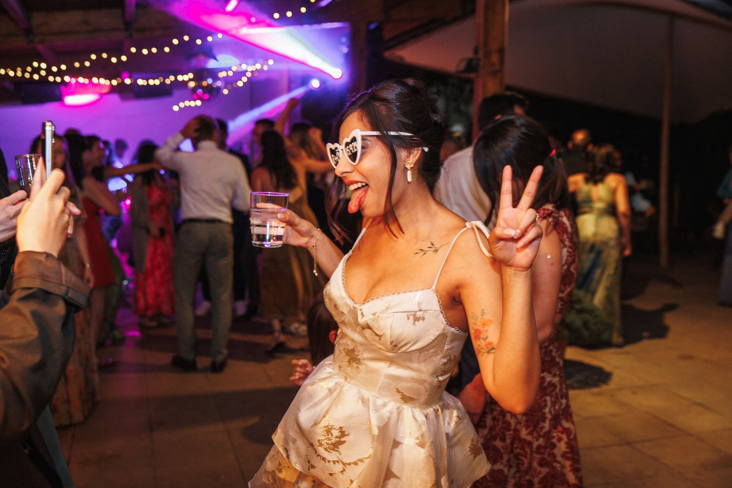 Bride in white floral dress and cat-eye sunglasses dancing and making peace sign at wedding reception at La Baronia, Barcelona