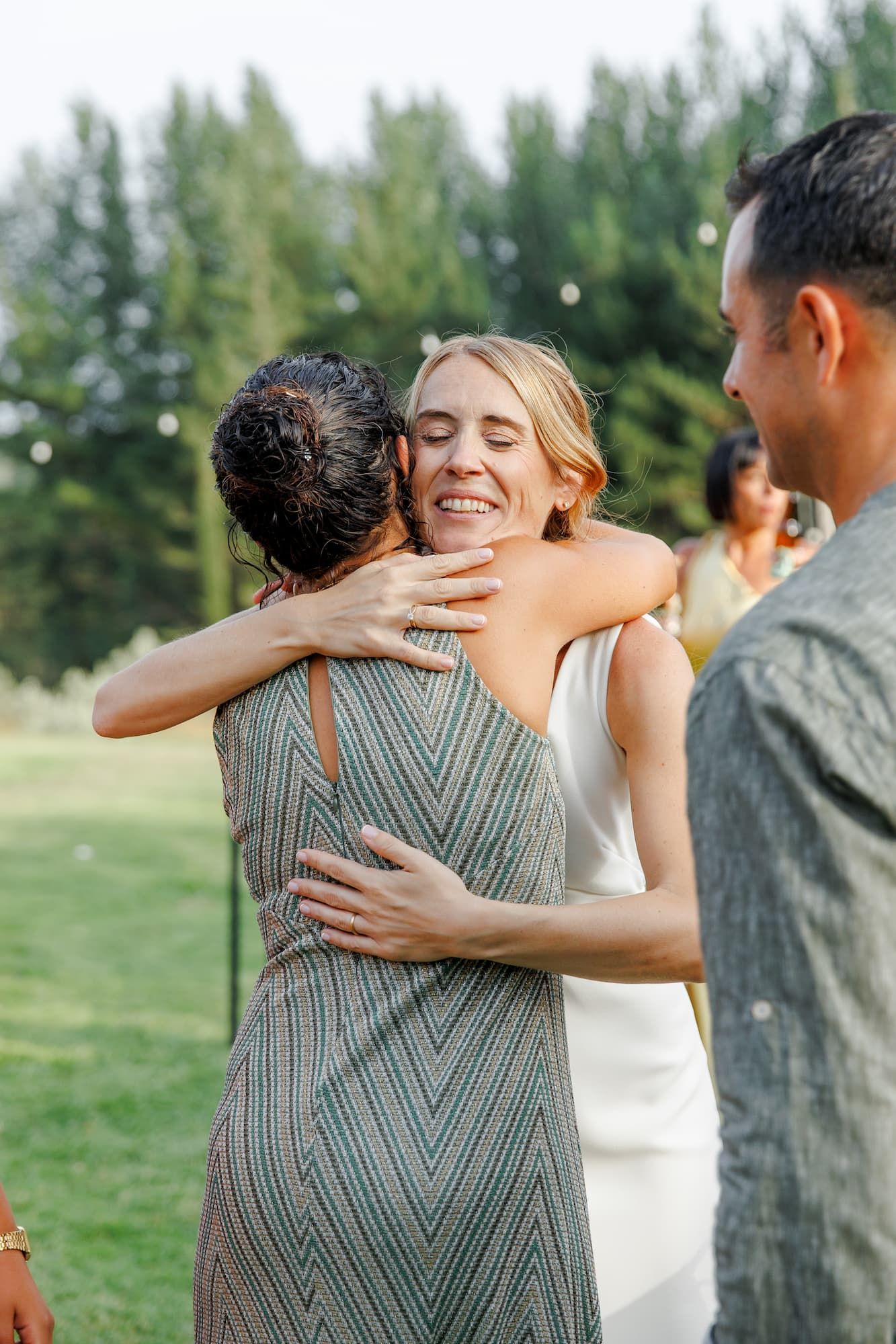 Bride in white dress joyfully hugging a guest in a green herringbone dress at an outdoor wedding reception.