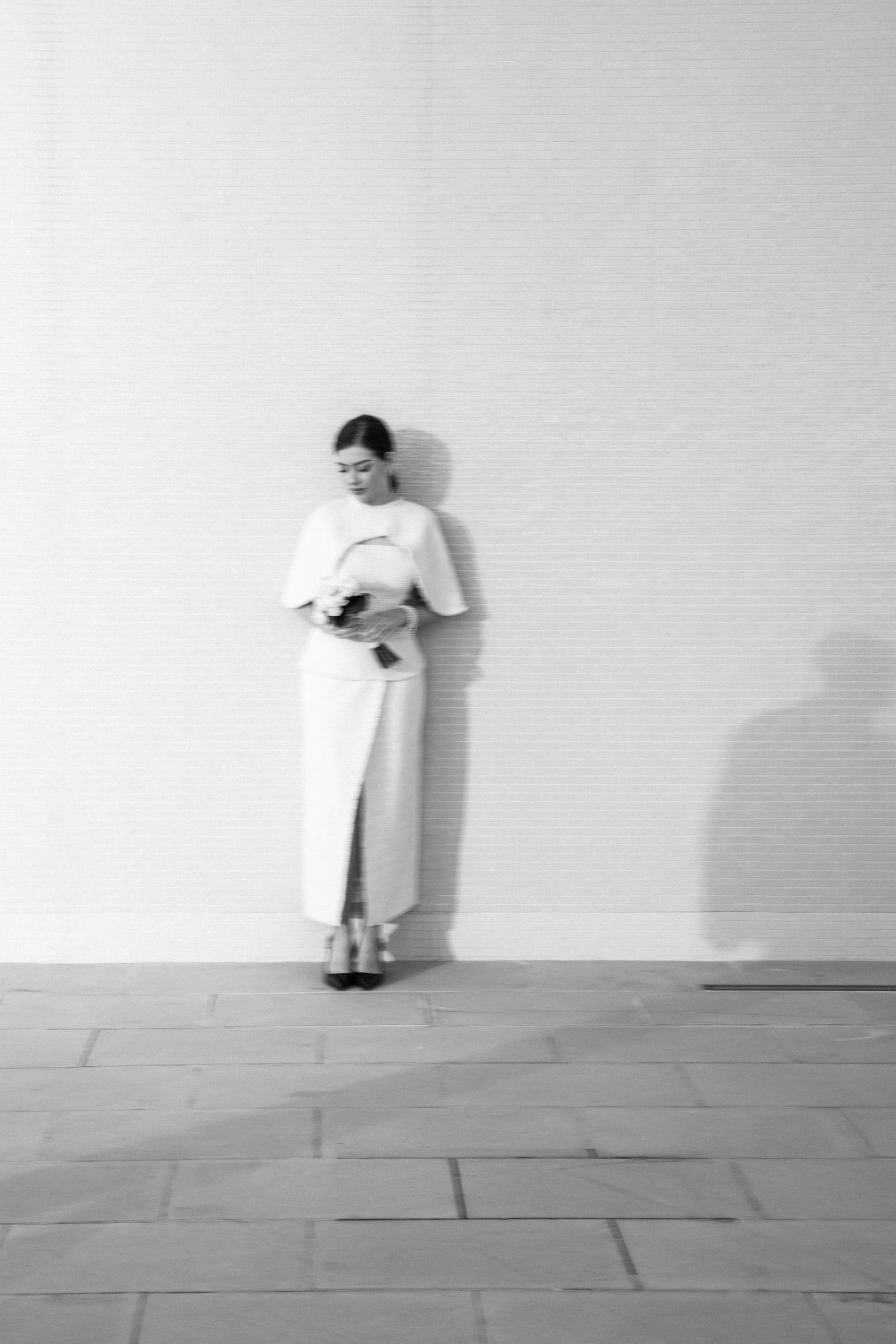 Black-and-white photo of a bride in a white dress, leaning against a wall, looking at her bouquet.