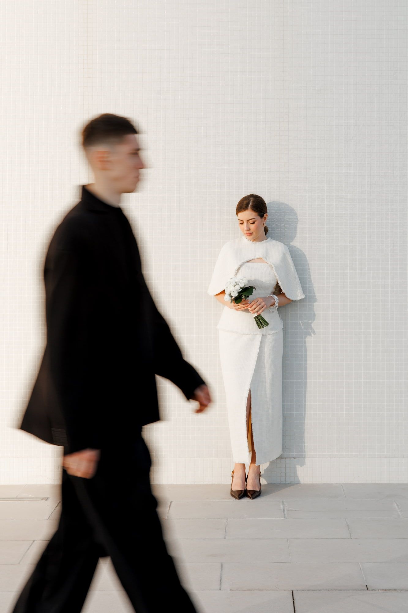 Bride in a white dress holds a bouquet against a white wall as the groom in a black suit walks past in motion blur.