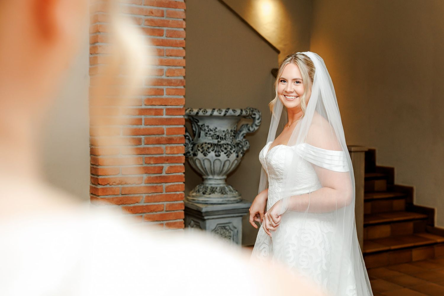 Smiling bride in white off-shoulder gown and veil stands near brick wall during wedding first look moment.