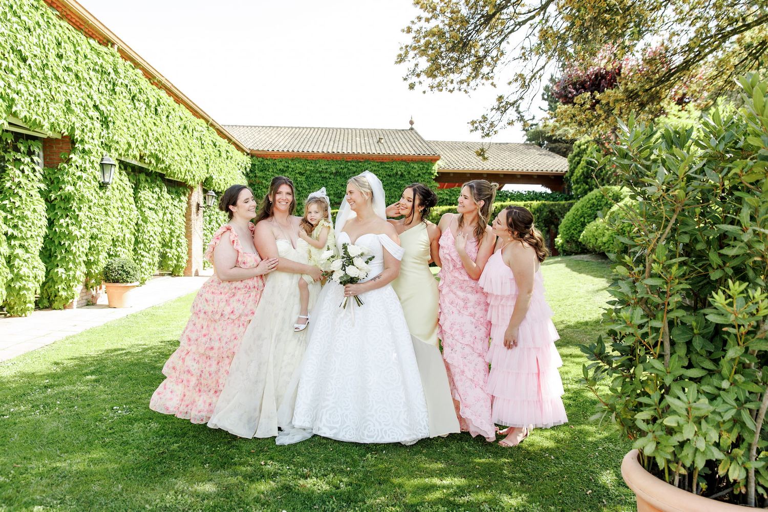 Bride in white gown surrounded by bridesmaids in pink dresses, posing outdoors at a garden wedding venue.