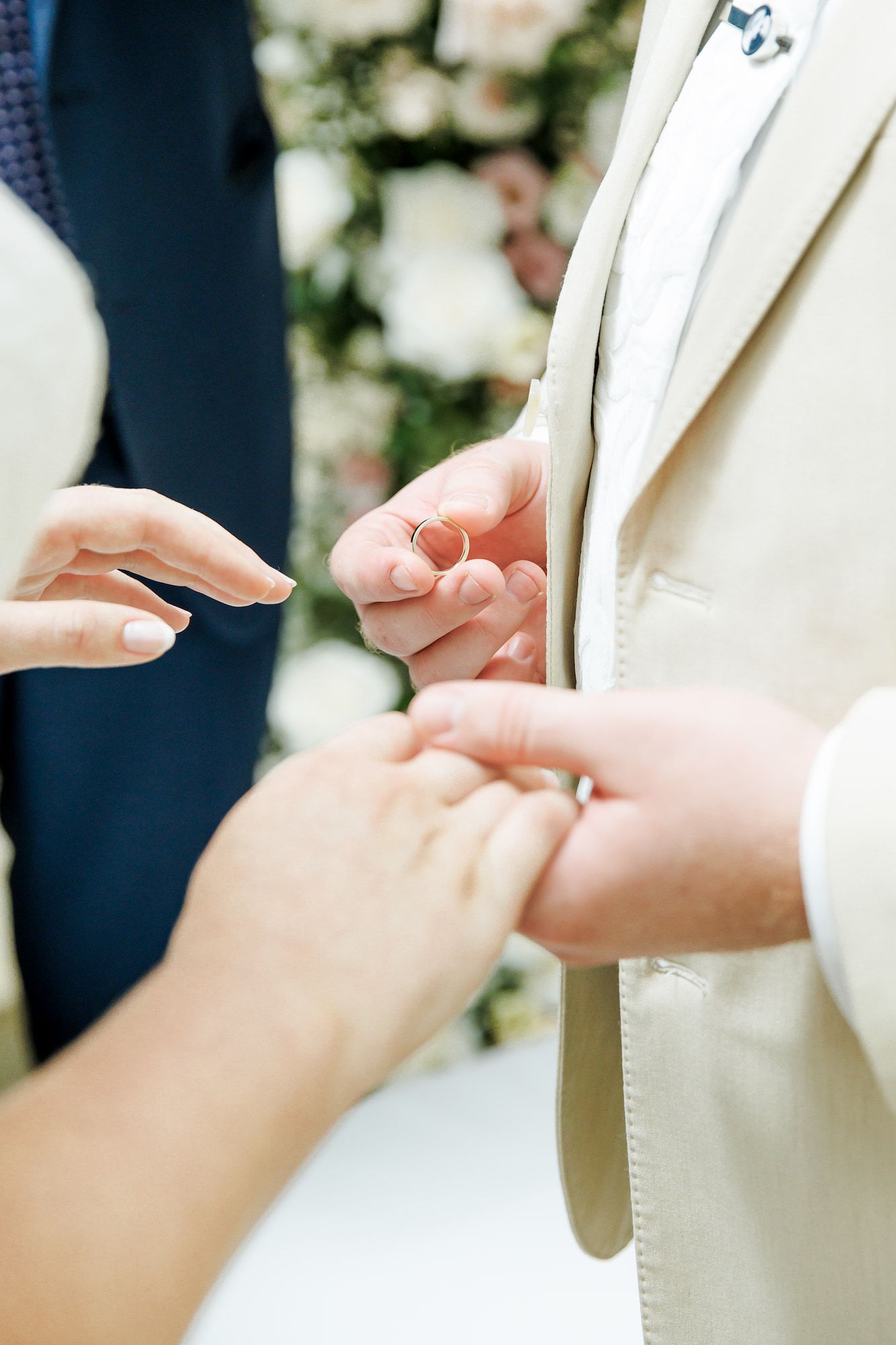 Groom placing a wedding ring on bride's finger during a ceremony with floral backdrop.