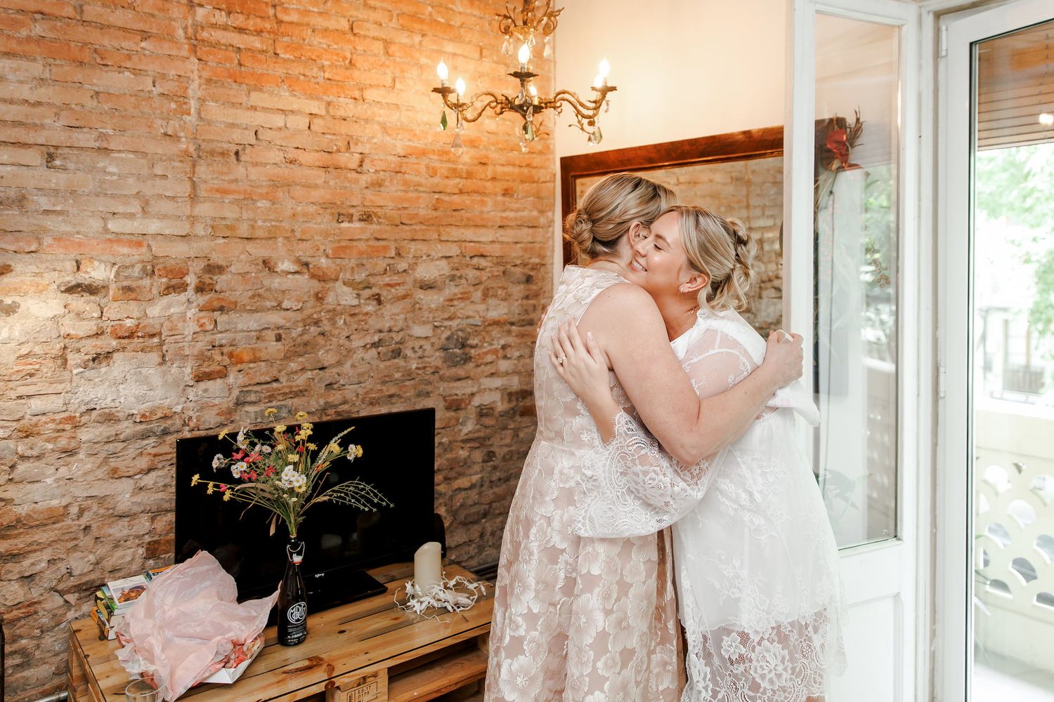 Two brides sharing an emotional hug in a rustic brick-walled room during wedding preparations.