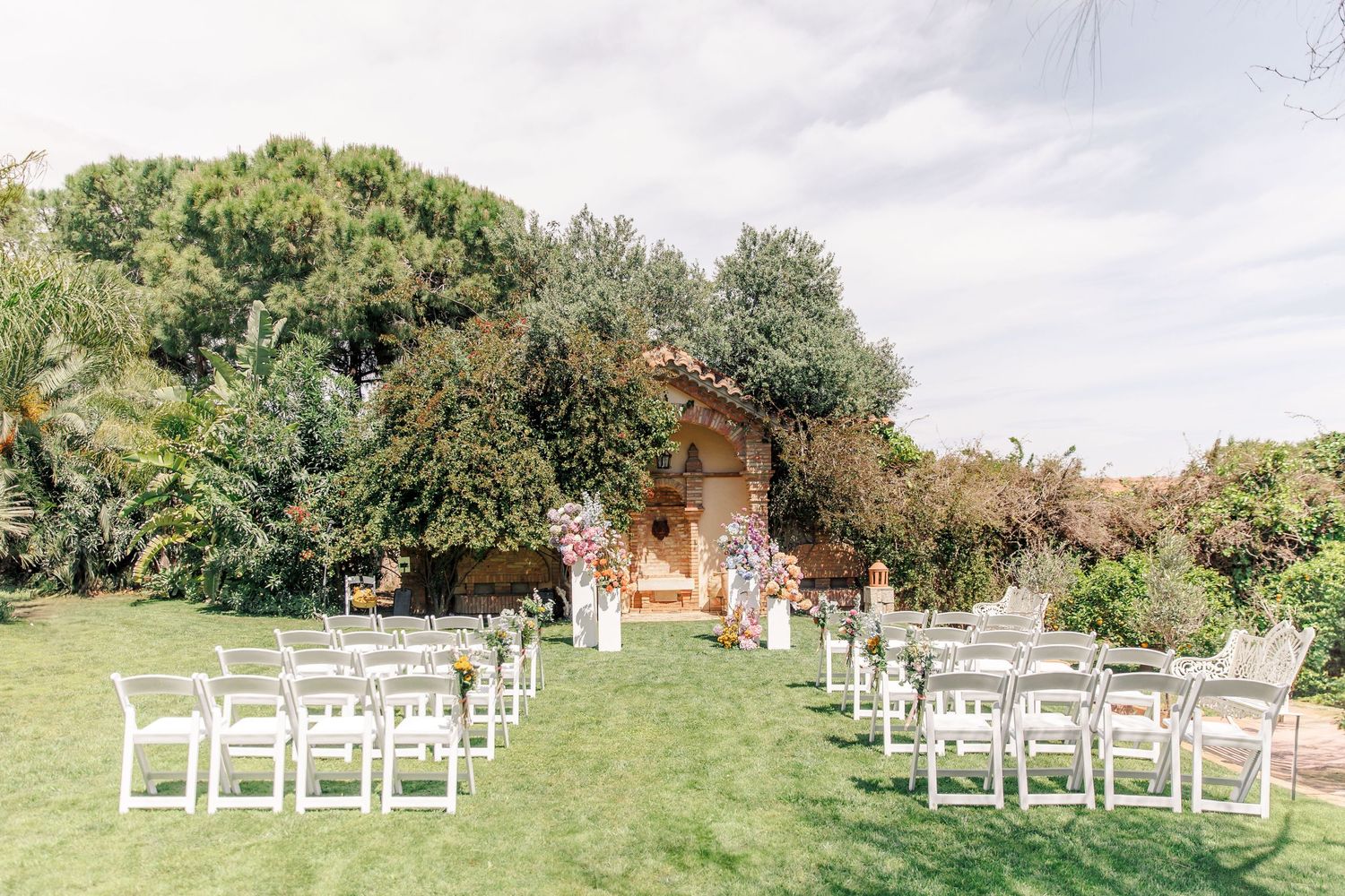 Outdoor wedding ceremony setup with white chairs on green lawn, floral arch, and rustic cottage backdrop.