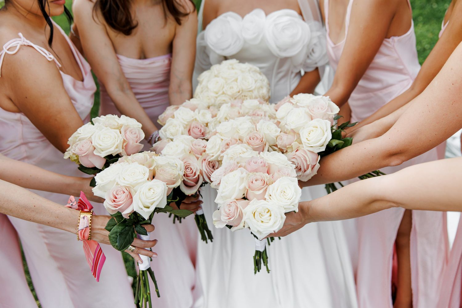Bride and bridesmaids in pink dresses holding bouquets of white and blush pink roses at a wedding.