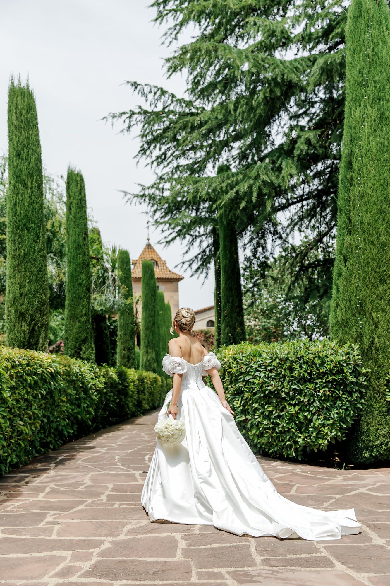 Bride in white off-shoulder gown with bouquet walks through elegant garden with cypress trees and historic building.